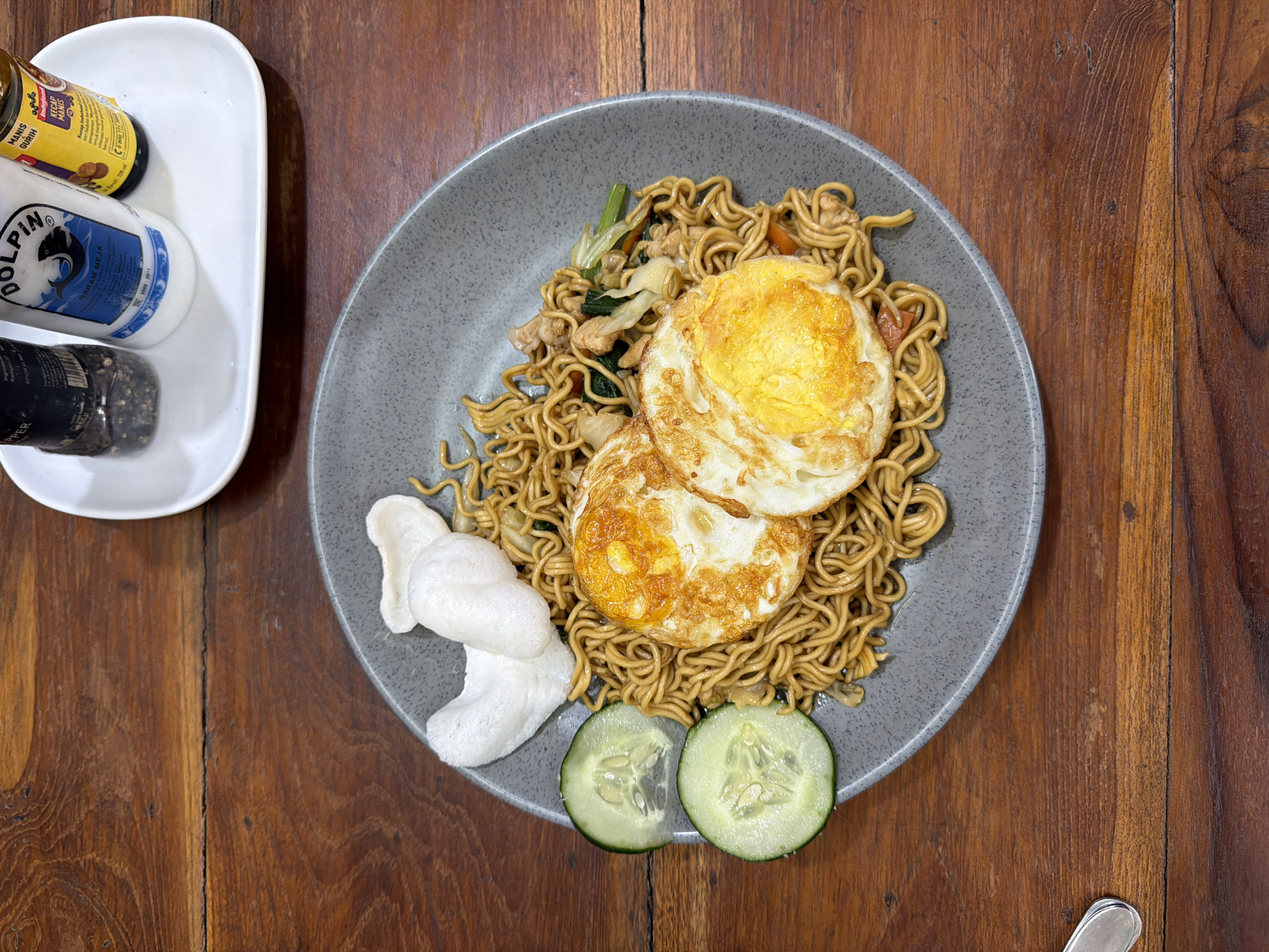 A top-down, medium shot depicts a plate of food on a wooden table, likely in Kuta, Indonesia, given the visible ingredients. The food consists of noodles topped with two fried eggs, garnished with two slices of cucumber and white crackers. To the left of the plate is a white tray holding a bottle of soy sauce, a salt shaker labeled "DOLPIN," and a pepper grinder. The table is made of dark wood. The plate is gray and speckled.