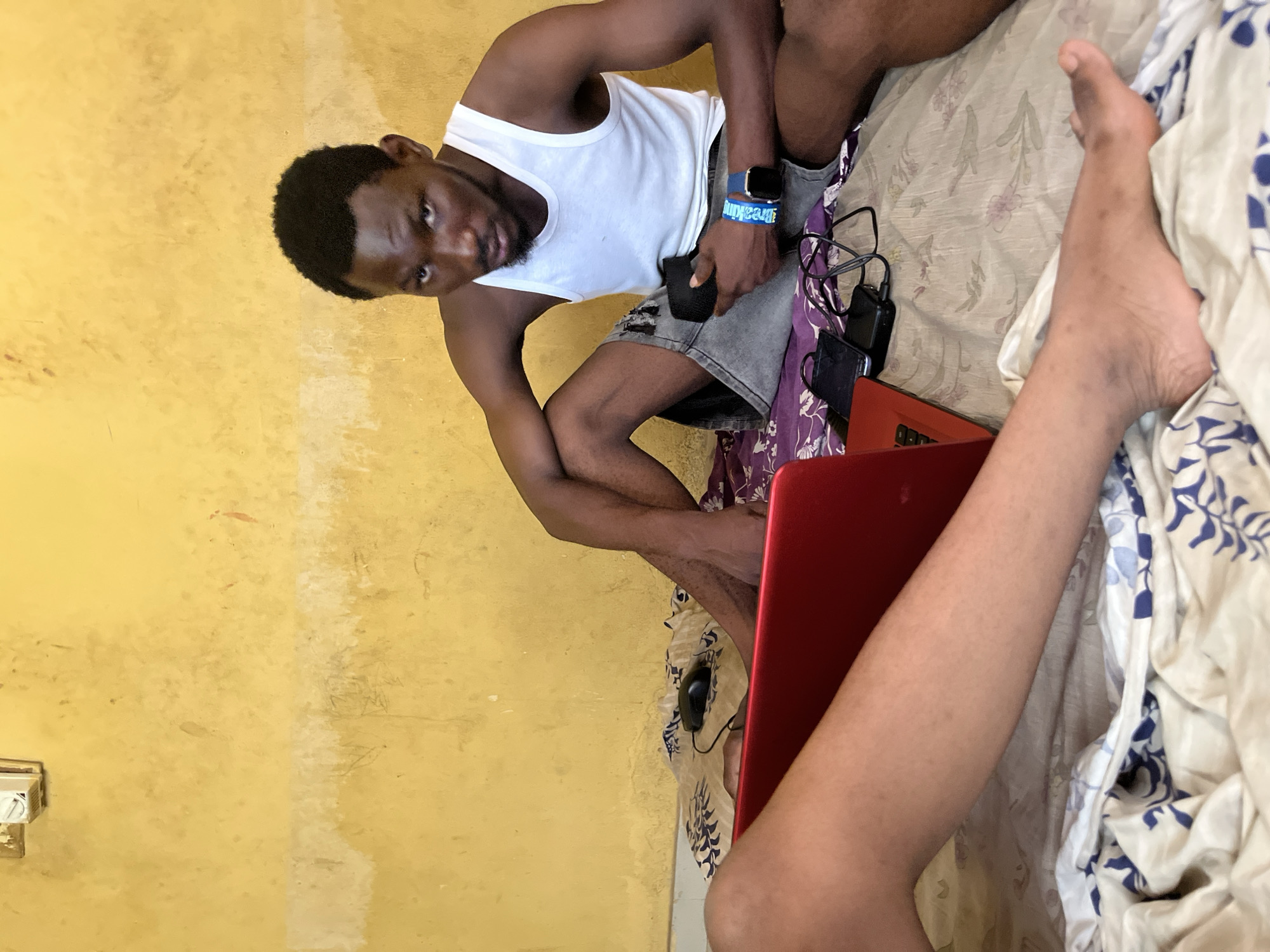 Two individuals are seated on a bed in a room with yellow, textured walls. The person in the foreground is partially visible from the waist down, their legs resting on a white and blue patterned duvet. A red laptop is positioned to their left. The other individual, a man, is seated further back, facing the camera. He is wearing a white tank top and dark denim shorts, and is holding a black object in his right hand. A blue smartwatch with a white band is visible on his left wrist. Various electronic devices, including a black mobile phone, a black power bank, and cables, are scattered on the bed between the individuals. A black computer mouse is also visible to the left of the laptop. The environment appears to be a domestic setting, possibly a bedroom.