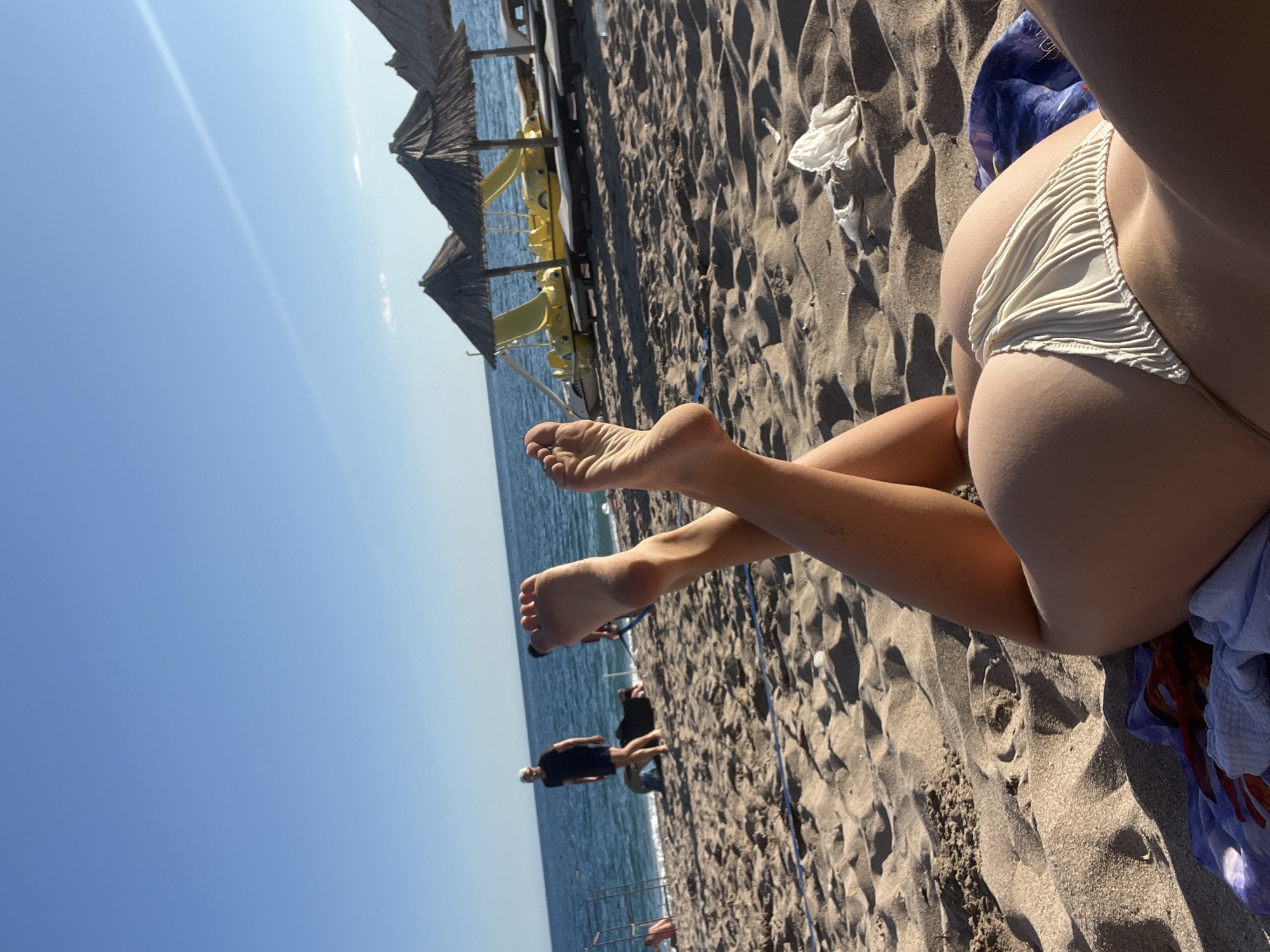 This image depicts a beach scene in Zankovići, Montenegro. In the foreground, a person lies on the sand, with their legs crossed and raised. They are wearing a light-colored bikini bottom. The sand is textured with ripples and footprints. In the mid-ground, there are several thatched umbrellas providing shade, and yellow pedal boats are visible behind them. The sea is calm and blue, meeting a clear sky. In the background, a man in a black t-shirt and shorts walks along the shoreline. Other people and beach structures are also present in the distance. The overall scene conveys a relaxed atmosphere on a sunny day at the beach.
