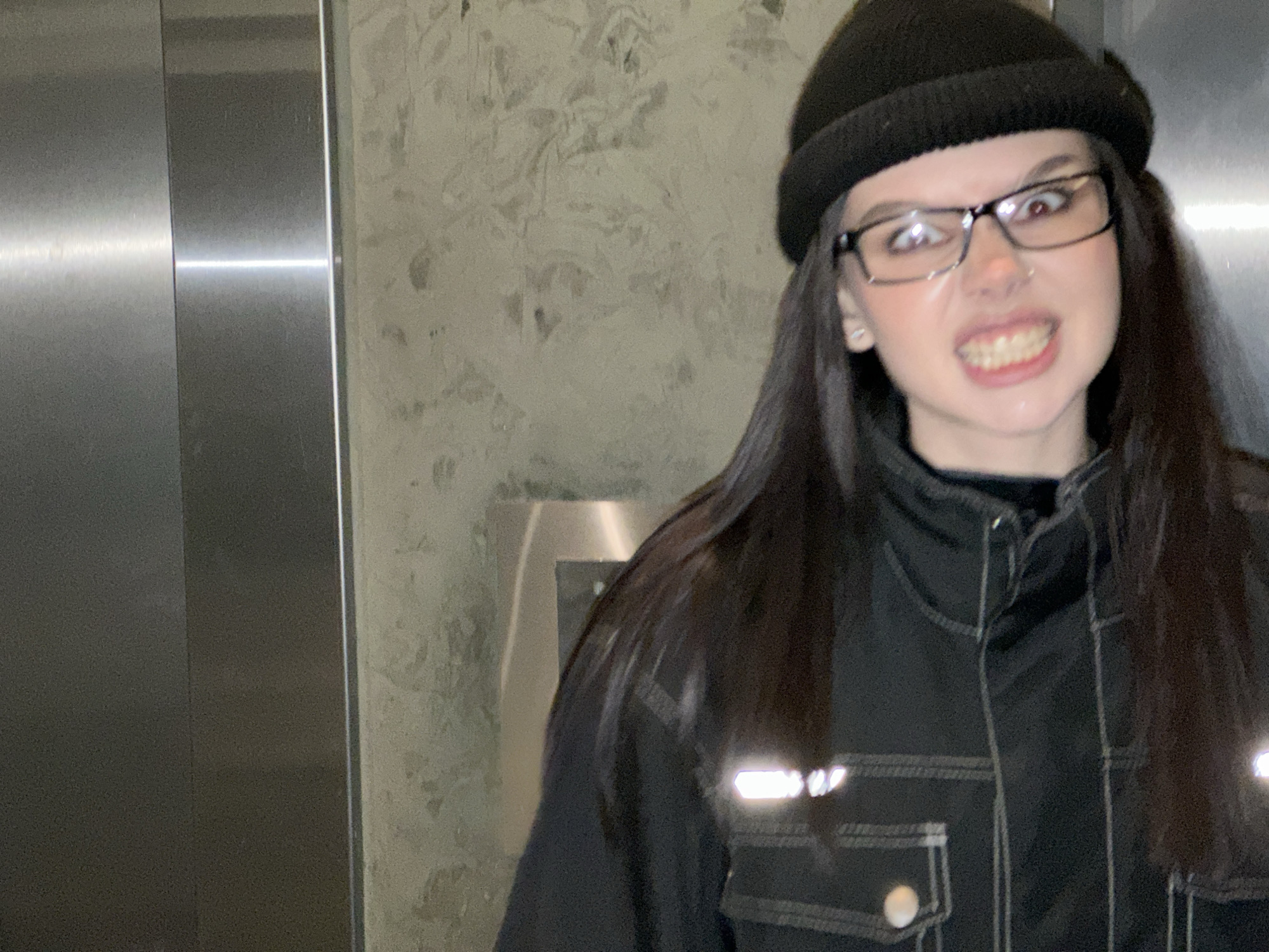 A person with long, dark hair, wearing a black beanie, glasses, and a dark jacket with reflective strips, is grimacing at the camera. The background includes a brushed metal surface, possibly an elevator door, and a textured wall. The location is likely an urban environment, but specific details to pinpoint the city are not available.