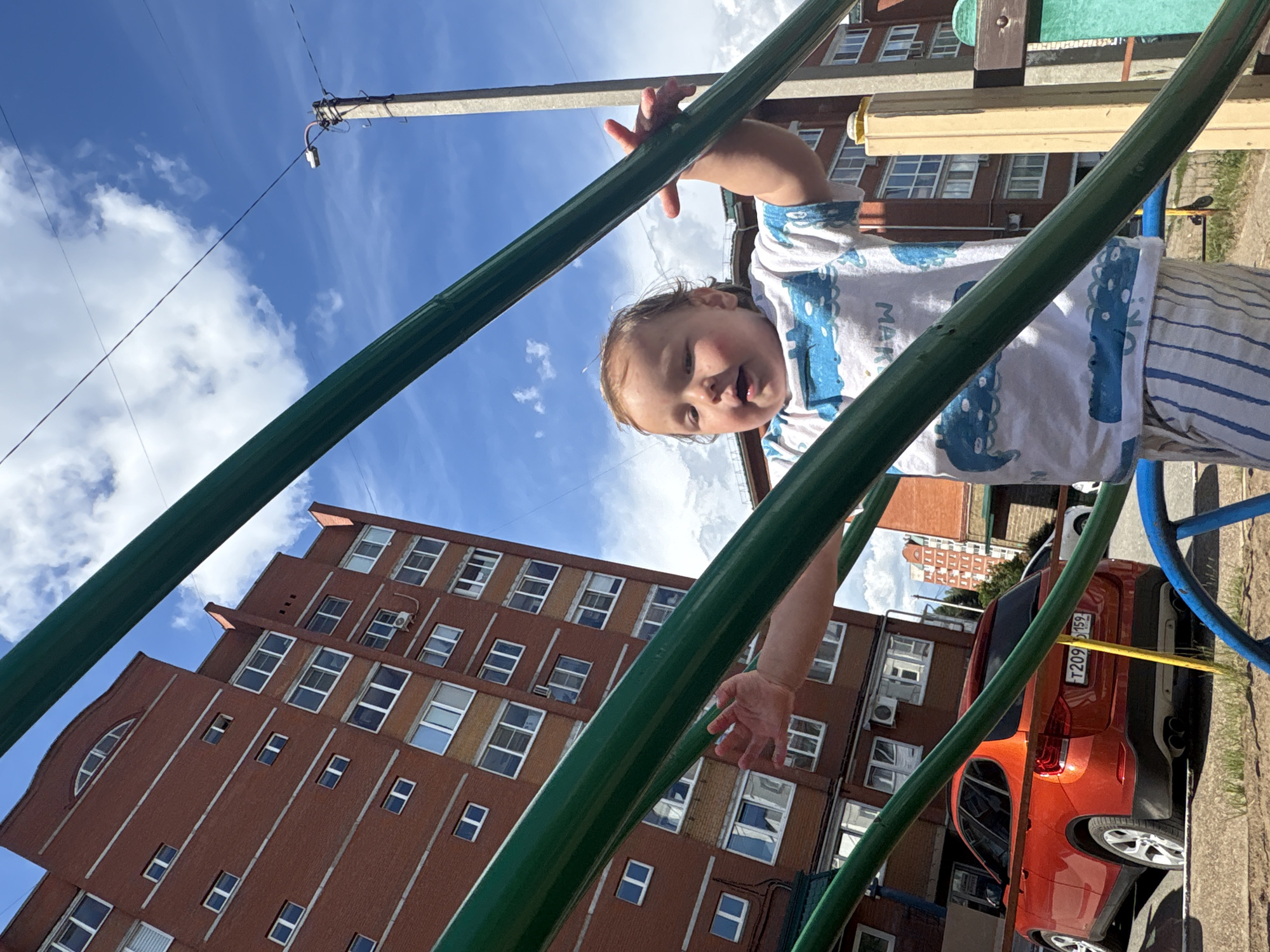 A low-angle shot captures a baby on a playground swing set, looking up at the sky. The baby is wearing a white t-shirt with a blue and white dinosaur print, and striped pants. Their face is tilted upwards, with an open mouth and eyes looking towards the camera. The background features a tall, brick apartment building, a blue sky with scattered white clouds, and a section of a street with a bright orange car parked. A utility pole with wires is also visible against the sky. The scene appears to be outdoors on a bright, sunny day. The image is a candid shot, capturing a playful moment of a child on a playground. There is text on the car's license plate, reading "T209". The location context is Chaikovsky, Russia.
