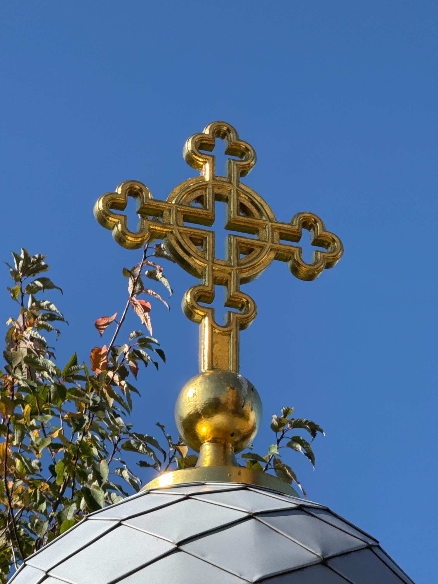 A golden cross stands atop a domed structure against a clear blue sky. The cross is ornate with cloverleaf-shaped ends. To the left of the cross, a tree with green and reddish-brown leaves is visible. The dome is covered in silver-colored, diamond-shaped shingles. The image appears to be taken on a sunny day. The location is not specified in the image.