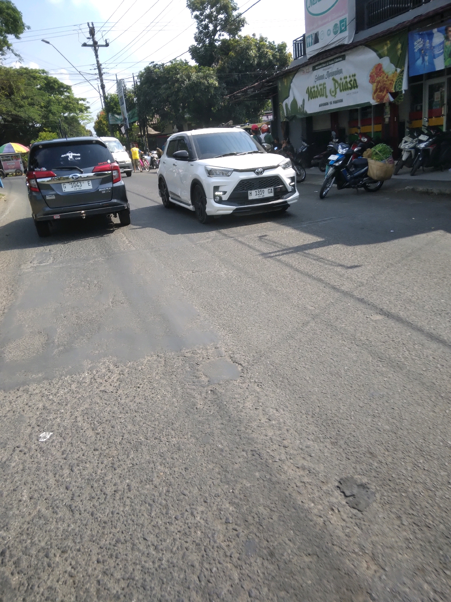 The image shows a street scene in Desa Pamijen, Kecamatan Sokaraja, Indonesia. The asphalt road is in poor condition with visible patching and potholes. A dark gray car with license plate G 1043 E is in the left lane, and a white SUV with license plate H 1319 GA is in the right lane. A street vendor's cart with an umbrella is partially visible on the left side of the road. In the background, a building with a banner that reads "Selamat Menunaikan Ibadah Puasa" is visible. Several motorcycles are parked in front of the building. Trees line the sides of the street, and utility poles with numerous wires are present. The sky is clear with some clouds.