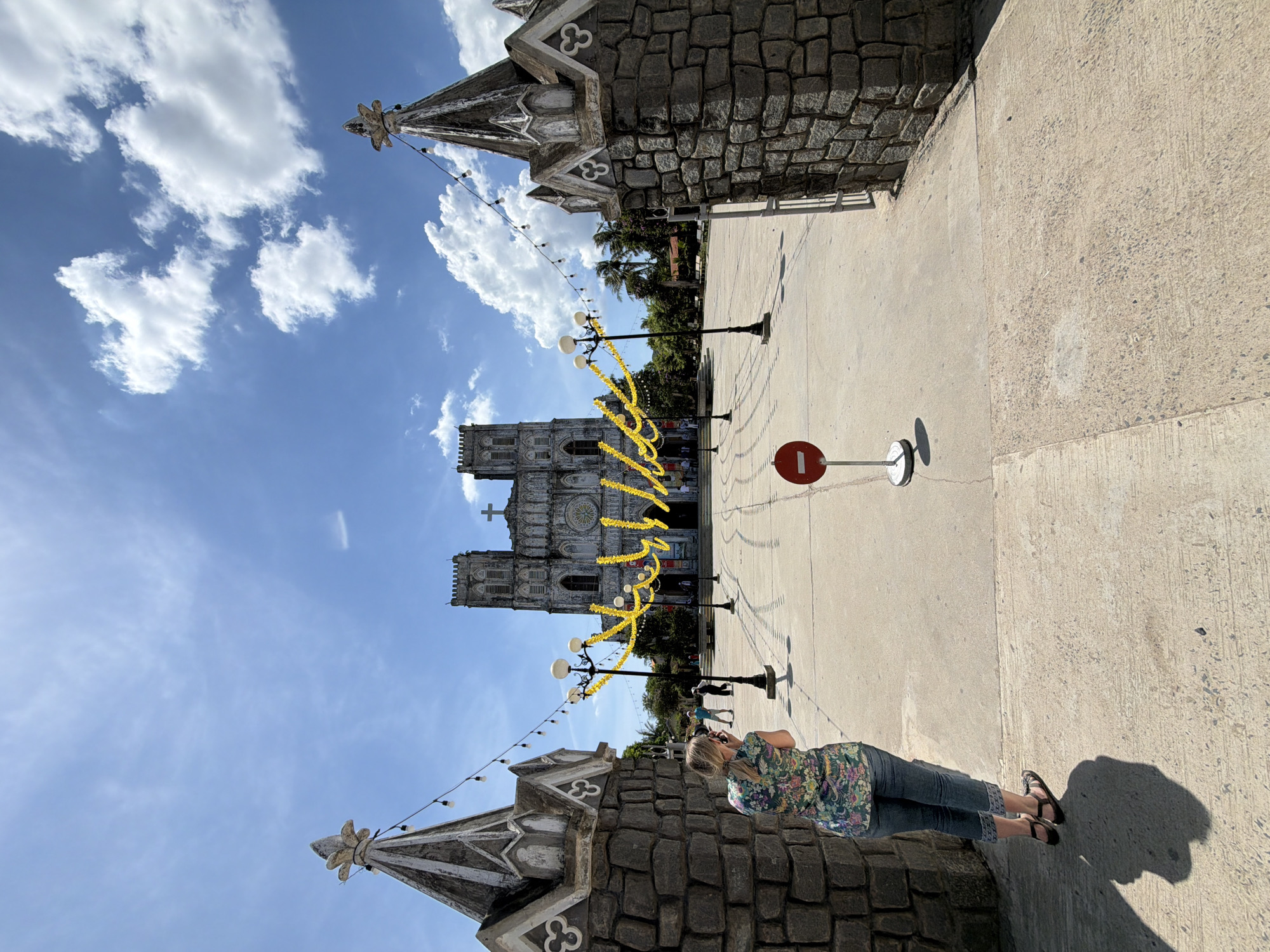 In front of the Saint Joseph Cathedral in the city of Hội Tín, Vietnam, a woman is seen from behind, standing on the courtyard and taking a picture of the cathedral. She is wearing a floral print shirt and blue jeans. The cathedral, an imposing structure with two bell towers and a large cross on its roof, is adorned with yellow garlands and string lights. The courtyard is paved with concrete, and a red and white "no entry" sign is visible in the foreground. The sky is blue with scattered white clouds, and palm trees can be seen in the background, suggesting a tropical climate. The scene is captured on a bright, sunny day.