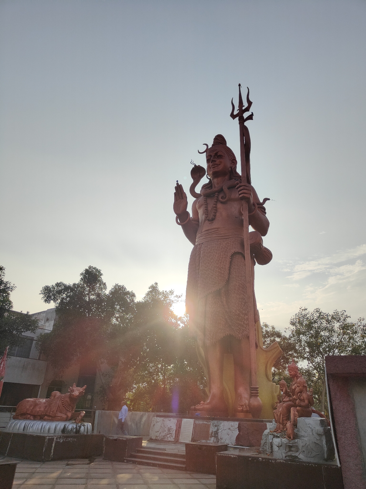 The image captures a low-angle, wide shot of the colossal statue of Lord Shiva at the Shiva Temple in Sector 29, Gurgaon, India. The sun is setting, casting a warm glow and lens flares across the scene, with trees and a building visible in the background. The statue, made of reddish-brown material, depicts Lord Shiva in a standing pose, holding a trident and a damaru, with snakes around his neck and arm. To the left of the main statue, a smaller statue of a bull is also visible. To the right, at the base of the main statue, there are smaller sculptures of deities seated on rocks. A person in a light-colored shirt walks in the middle ground, appearing to observe the statues. The ground in the foreground is paved.