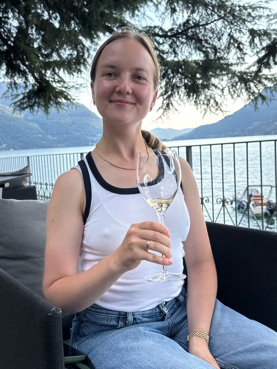 A woman is sitting on a dark sofa outdoors, holding a glass of white wine. She is wearing a white tank top with black trim and blue jeans. She has a ring on her left ring finger and a bracelet on her left wrist. In the background, there is a body of water with mountains and trees. The woman is smiling at the camera. The scene appears to be a restaurant or bar overlooking a lake in Pognana Lario, Italy.