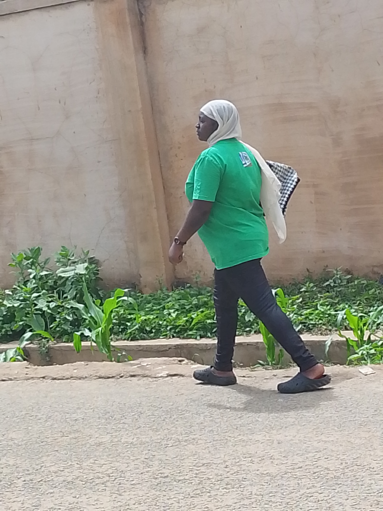 A woman is walking along a street in Jos, Nigeria. She is wearing a white headscarf, a green T-shirt, black pants, and black slip-on shoes. She is carrying a black and white checkered bag over her shoulder. She is walking on a paved street with a raised concrete barrier on the left side of the frame. Beyond the barrier, there is a line of green plants. Behind the plants is a concrete wall. The scene is lit by natural daylight.