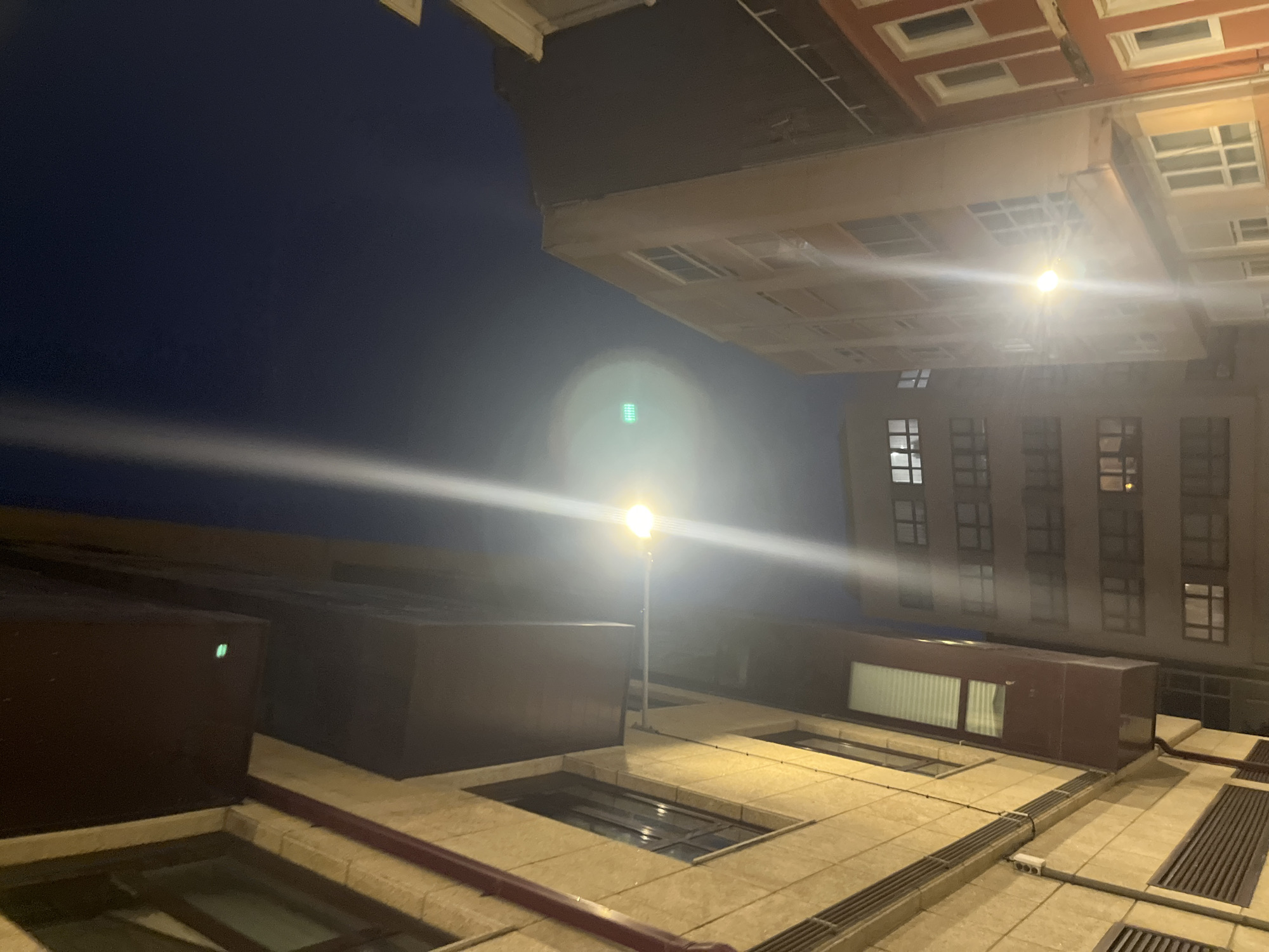 The image shows an urban scene at night, likely taken in A Coruña, Spain, based on architectural styles and lighting. The foreground features building rooftops with ventilation grates and a reddish-brown pipe or cable running along the edge. A street lamp stands prominently in the middle, casting a bright light and glare. In the background, buildings with multiple windows are visible, partially obscured by the light from the street lamp. The sky is dark, suggesting a time after dusk.