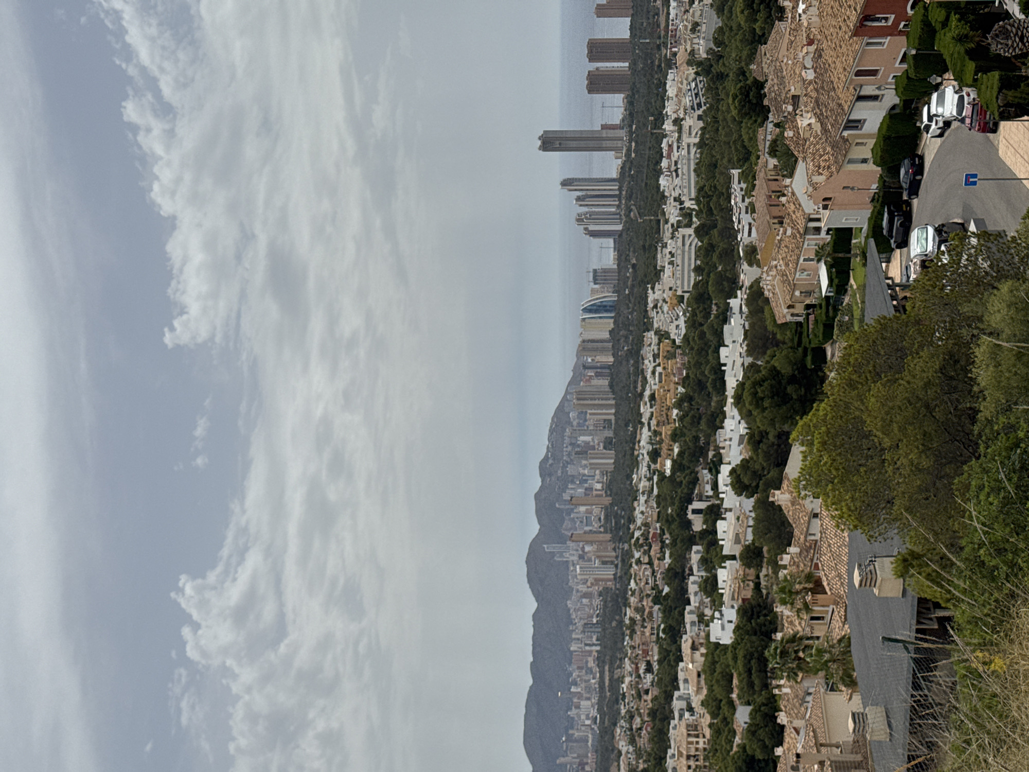 This is a daytime, high-angle, wide shot of the city of Urbanització Golf Badia, Spain. The sky is overcast with white and grey clouds. In the distance, a range of dark, undulating mountains forms the horizon. Beyond the mountains, the sea is visible, appearing as a pale grey expanse.
The middle ground of the image is dominated by a densely packed cityscape featuring numerous high-rise buildings, many of which are residential towers. The buildings vary in height and architectural style, with some appearing modern and sleek, while others have a more traditional design. Some of these buildings are located along the coastline.
In the foreground, the scene transitions to a residential neighborhood characterized by smaller, terracotta-roofed houses with light-colored facades. These houses are surrounded by lush green trees and manicured hedges. A paved road curves through the neighborhood, with several cars parked along its edge, including a black SUV, a black sedan, a white sedan, and a white SUV. A street sign with a blue and white symbol is visible on the side of the road. The foreground also shows areas of dry grass and other vegetation, suggesting a hilly terrain. The overall impression is of a sprawling urban and suburban environment set against a natural mountainous backdrop.
