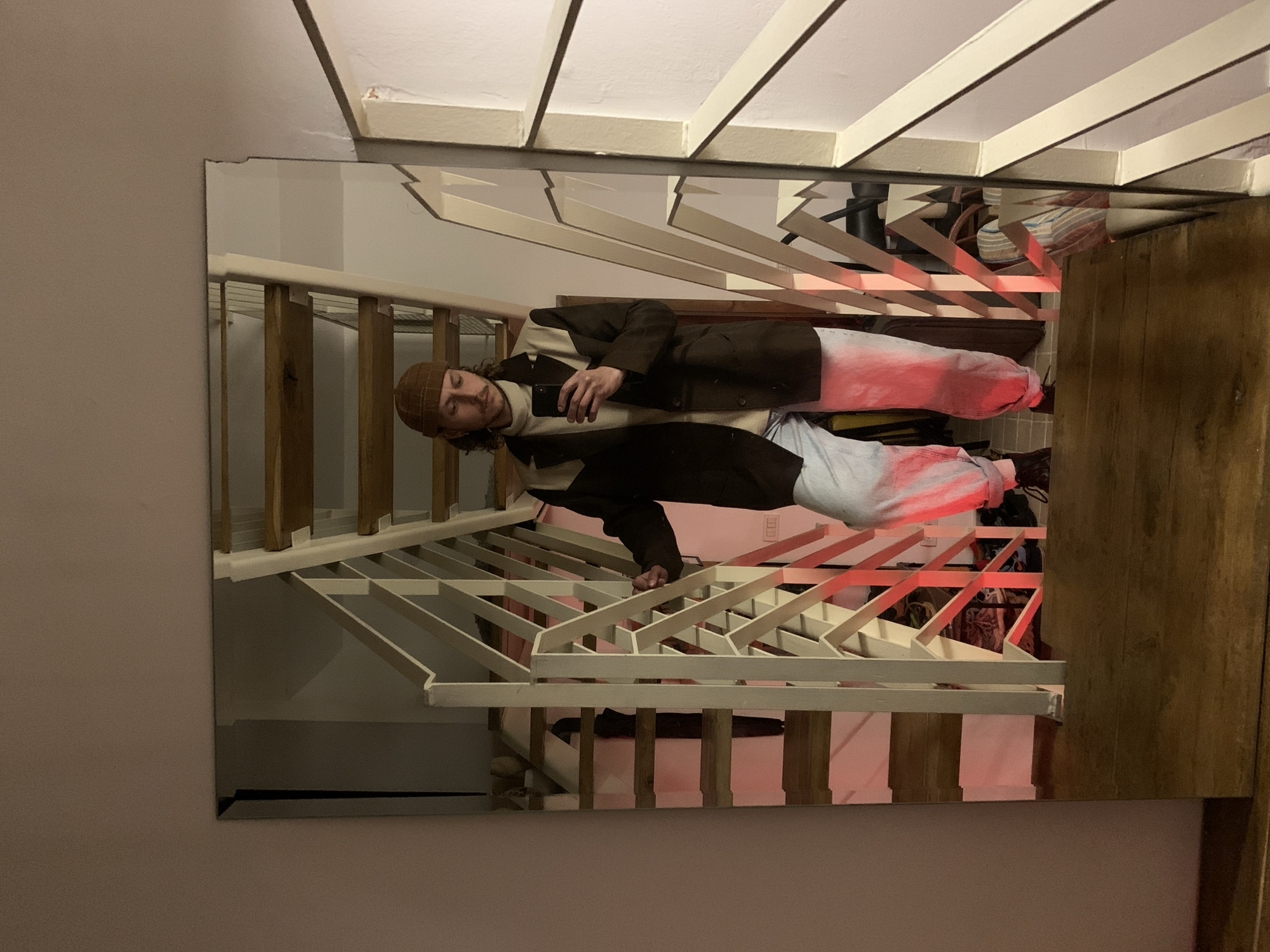 An indoor mirror selfie captures a male individual positioned on a wooden landing at the top of a staircase within a residential setting in Tel-Aviv, Israel. The scene is characterized by dim ambient lighting, with a distinct red glow cast upon the lower section of the staircase. The individual is a male with a mustache and dark, wavy hair, wearing a light brown beanie, a cream-colored turtleneck sweater, a dark brown blazer with cream-colored shoulder panels, light blue cuffed trousers, and dark footwear. He holds a black smartphone in his extended right hand, capturing his reflection, while his left hand rests on the white, geometrically patterned metal railing of the wooden staircase. The mirror reflects the descending staircase, beneath which various indistinct objects, including a dark elongated item and a patterned rug, are visible. A light-colored wall with an electrical outlet featuring red inserts is also reflected at the base of the stairs. A plain, light beige wall frames the mirror on its left and top edges.