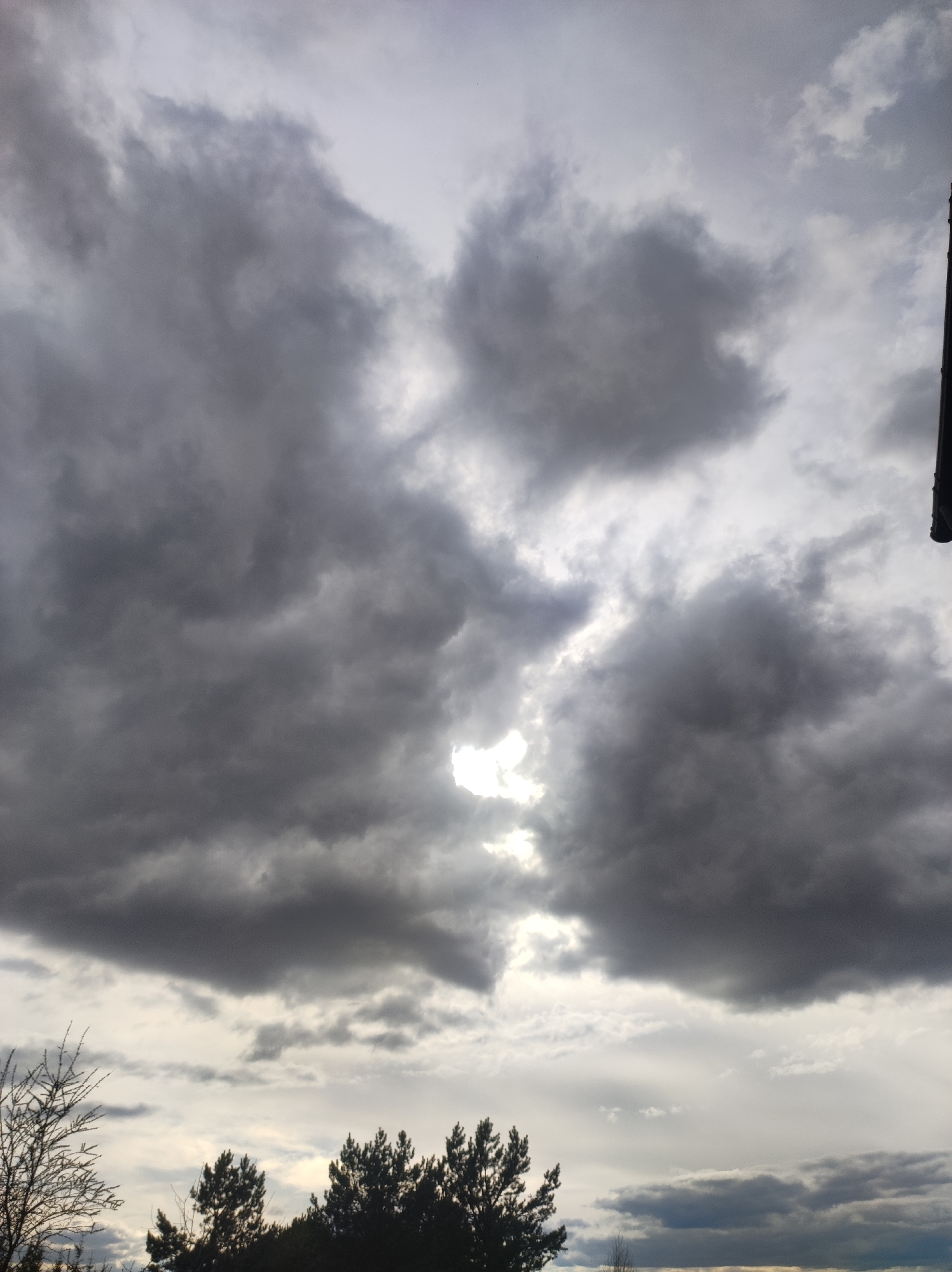 The image shows a cloudy sky with a small break in the clouds allowing some sunlight to shine through. Below the clouds, there are trees in silhouette. A portion of a dark structure is visible in the upper right corner. The location in the city of Фоки, Russia, cannot be confirmed from the image.