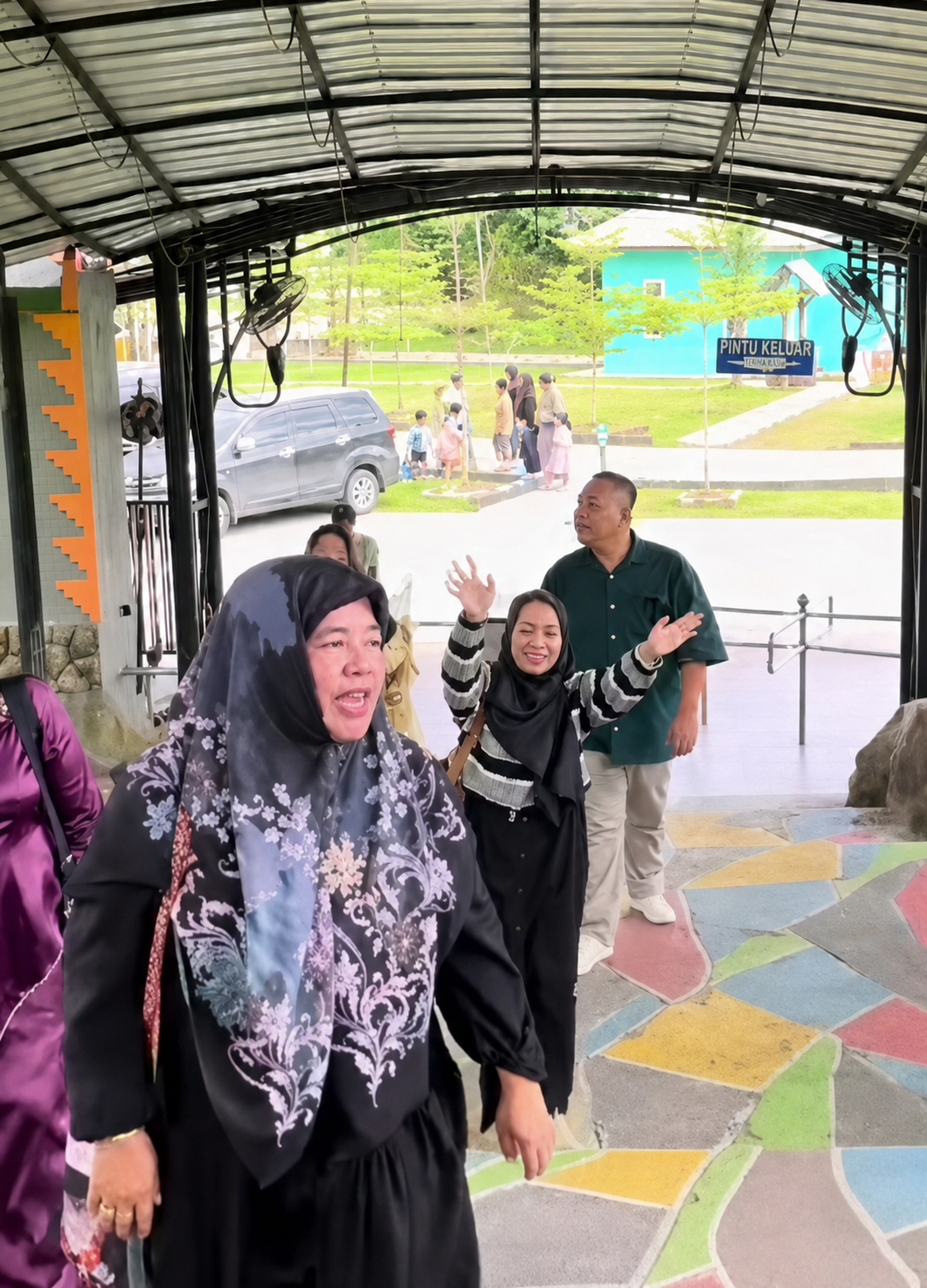 This image captures a group of people gathered under a covered walkway. In the foreground, a woman wearing a black hijab with a floral pattern is looking to her left and speaking animatedly. To her right, another woman in a black and white striped top and black hijab is smiling and waving with both hands. Behind them, a man in a dark green collared shirt and light-colored pants stands looking forward. Further back, a group of children and adults are visible in the background, with a turquoise building and trees suggesting an outdoor location. A sign reading "PINTU KELUAR" (EXIT) is also visible in the background. The floor of the walkway is decorated with colorful geometric shapes. The scene appears to be in a public space, possibly a park or an attraction in Eampat Kwalamencirim, Indonesia.