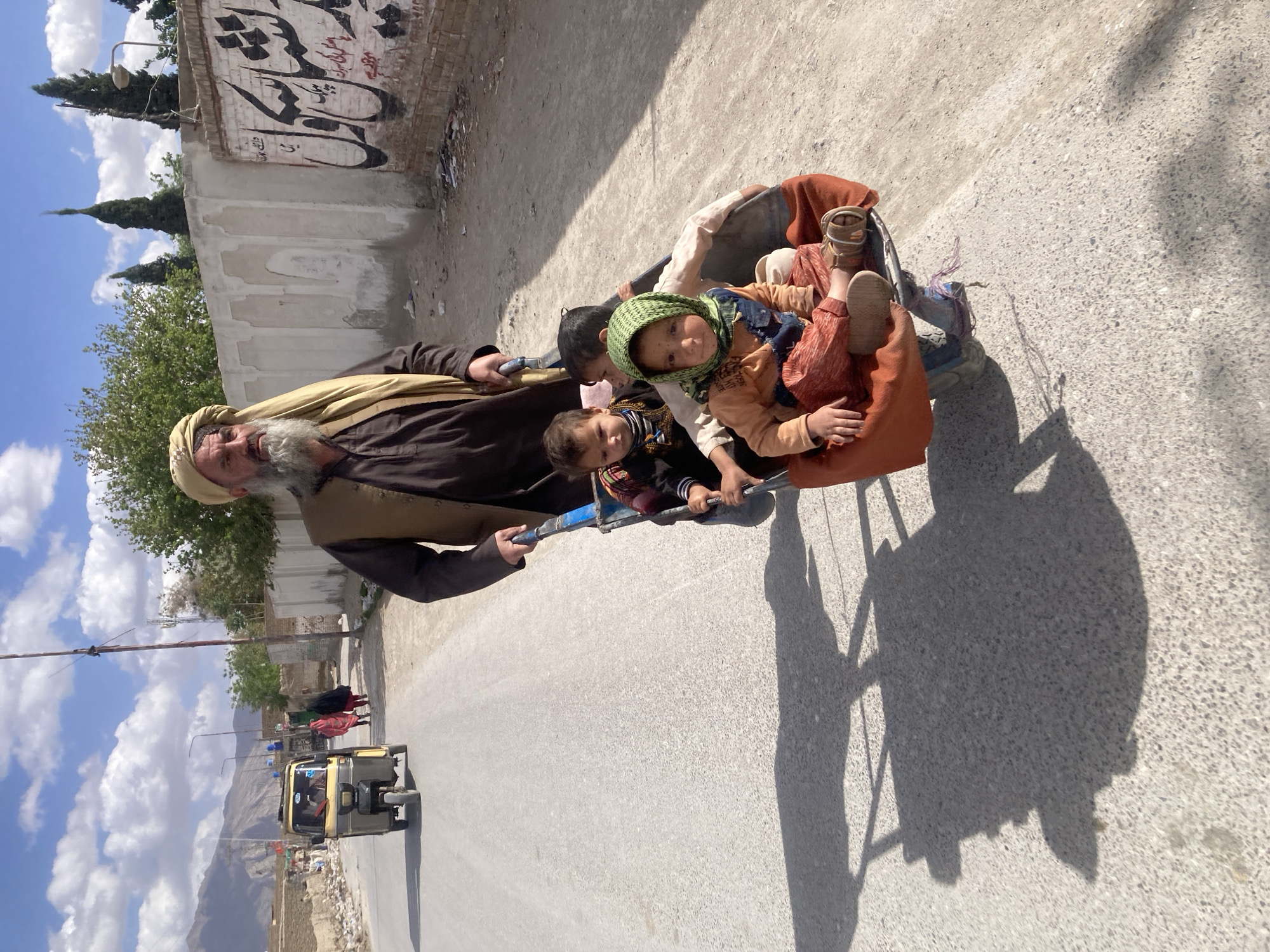 A man with a long white beard and a tan turban is pulling a blue wheelbarrow filled with several children down a paved road. The man is wearing a brown coat over a lighter brown vest. The children in the wheelbarrow are of varying ages and are wearing different colored clothing.
The road is lined with buildings and a wall covered in graffiti. In the background, there are mountains and a blue sky with clouds. A yellow auto rickshaw is driving in the opposite direction.
The location is Quetta City Tehsil, Pakistan.