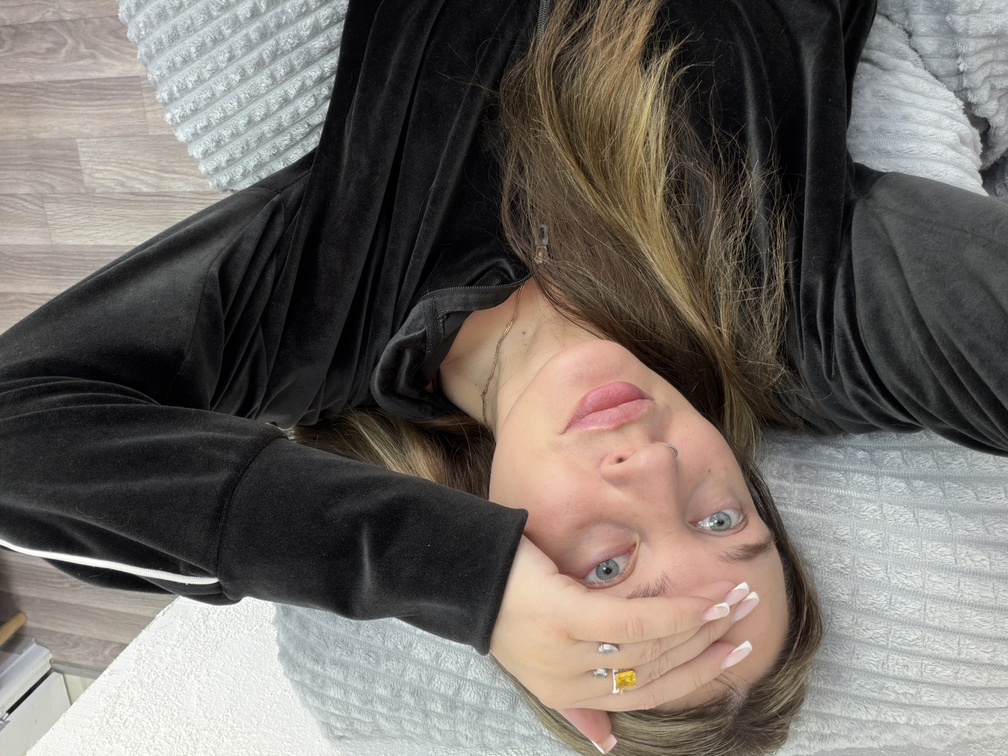 A medium-shot, angled portrait shows a woman lying on a white quilted surface, presumed to be a bed or treatment table. She is wearing a black zip-up jacket and has highlighted brown hair. Her left hand, adorned with a prominent yellow gemstone ring and a silver ring, is partially covering her face. The background includes a patterned gray pillow and a light-colored wooden surface, possibly flooring. There is no explicit evidence to suggest the location of the image is in Chaikovsky, Russia.