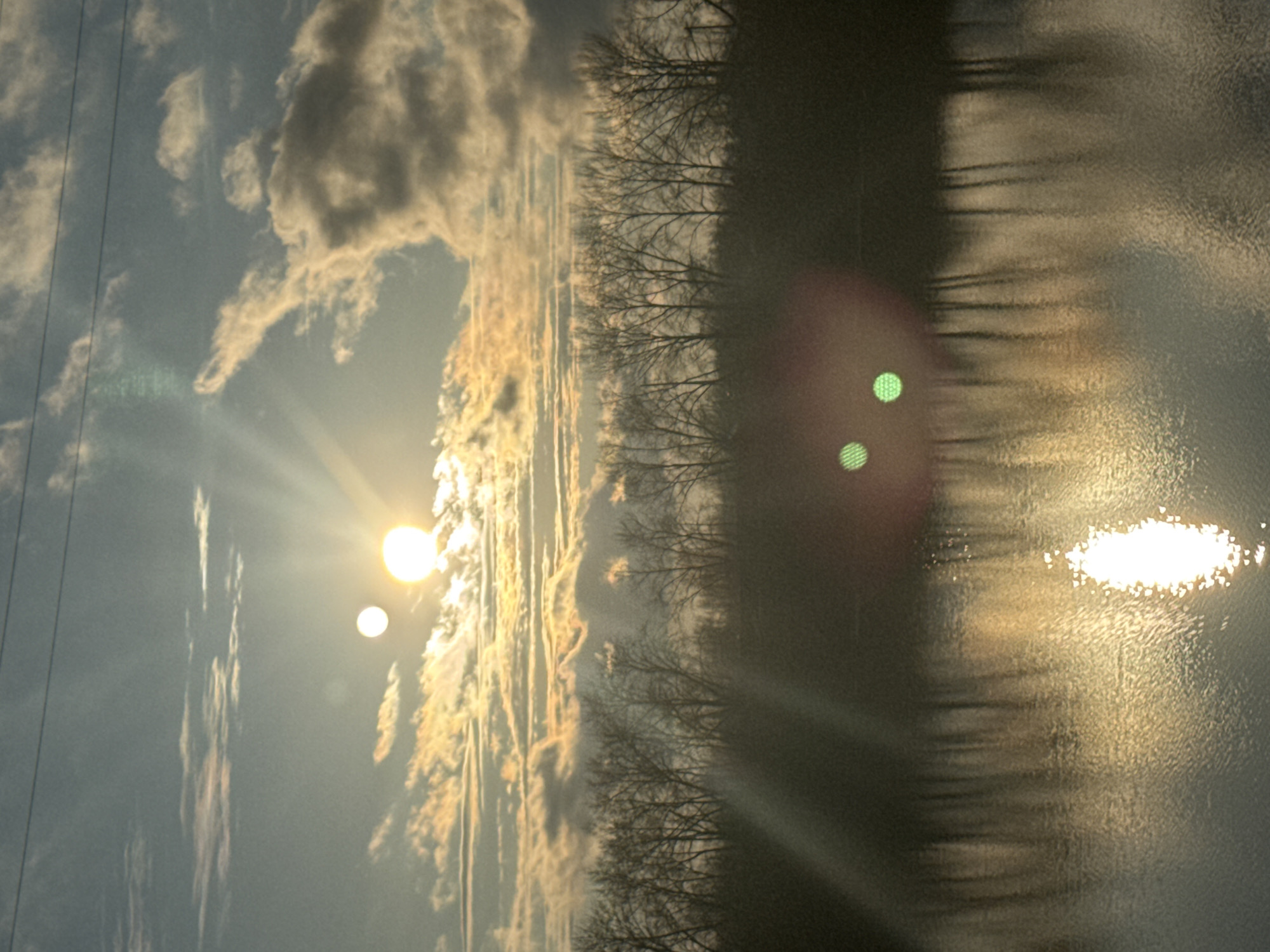 The image, when correctly oriented in landscape view, depicts a tranquil natural scene, likely a sunrise or sunset, in Chaikovsky, Russia. A bright, low-lying sun dominates the left side of the frame, emitting strong sun rays and creating a brilliant golden reflection across the surface of a body of water, possibly a river or lake.
The sky is partly cloudy, with some clouds appearing illuminated by the warm light, exhibiting shades of golden yellow and orange against a cooler blue-gray background. A prominent horizon line is formed by a row of bare, deciduous trees, silhouetted darkly against the brighter sky and water. These leafless trees suggest the image was captured during a colder season, such as winter or early spring/late autumn. Faint power lines are visible in the upper left portion of the sky.
The water surface appears textured, with shimmering patterns reflecting the intense light. Several photographic artifacts are noticeable, including distinct green circular lens flares and a larger reddish-orange glow in the lower right area of the image. There are no people visible, nor is there any discernible text.