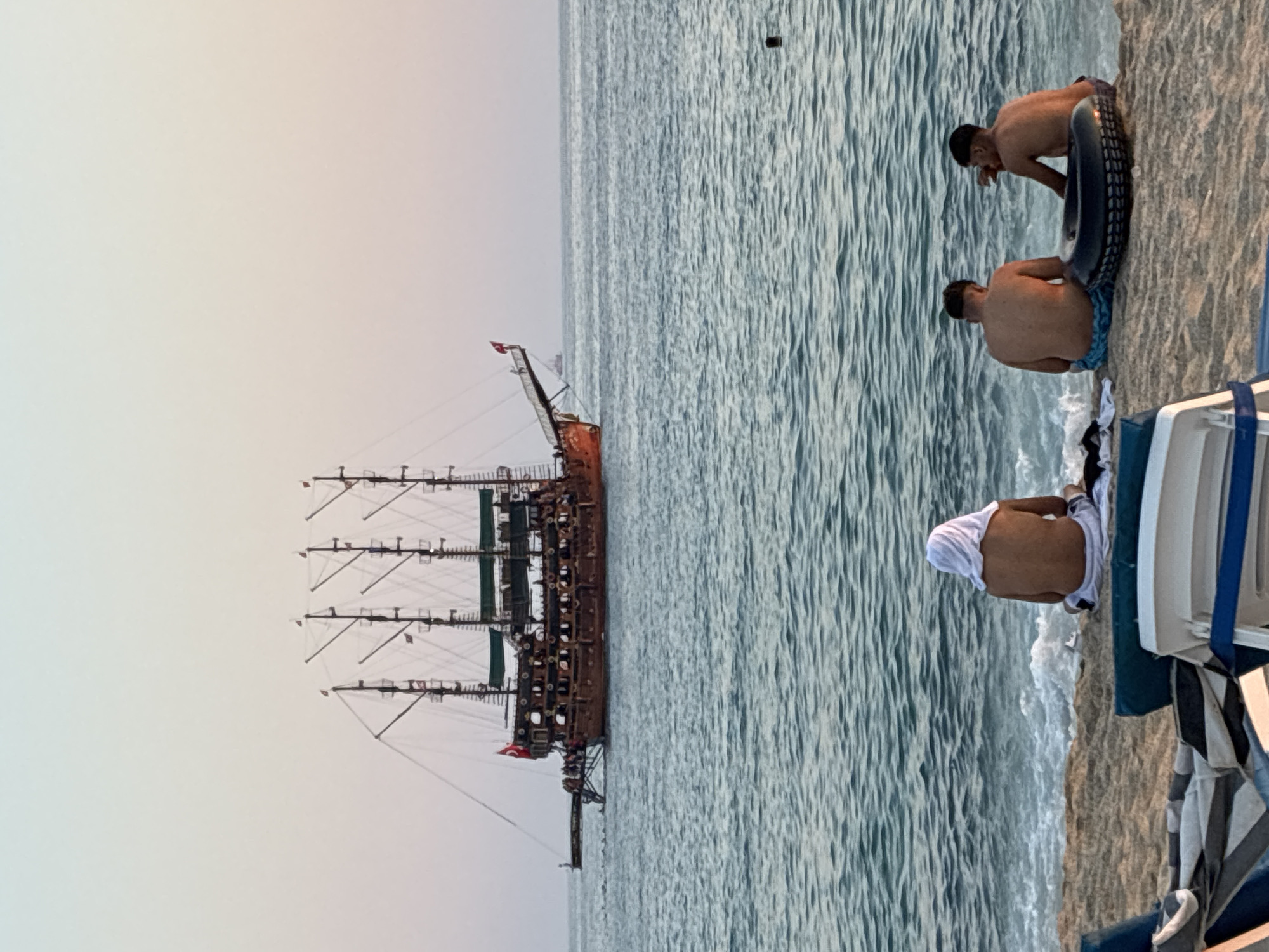 The image depicts a beach scene in Alanya, Turkey, with a large, multi-masted pirate-style ship sailing on the turquoise sea in the background. The sky above is a pale gradient of blue and pink, suggesting either sunrise or sunset. In the foreground, three young men are seated on the sandy beach, facing the water. The man on the left is wearing a white towel draped over his head and shoulders. The other two men are in swimwear, with a black inflatable ring placed between them. Waves are gently lapping at the shore. Beach chairs are visible in the very bottom left and center of the frame.