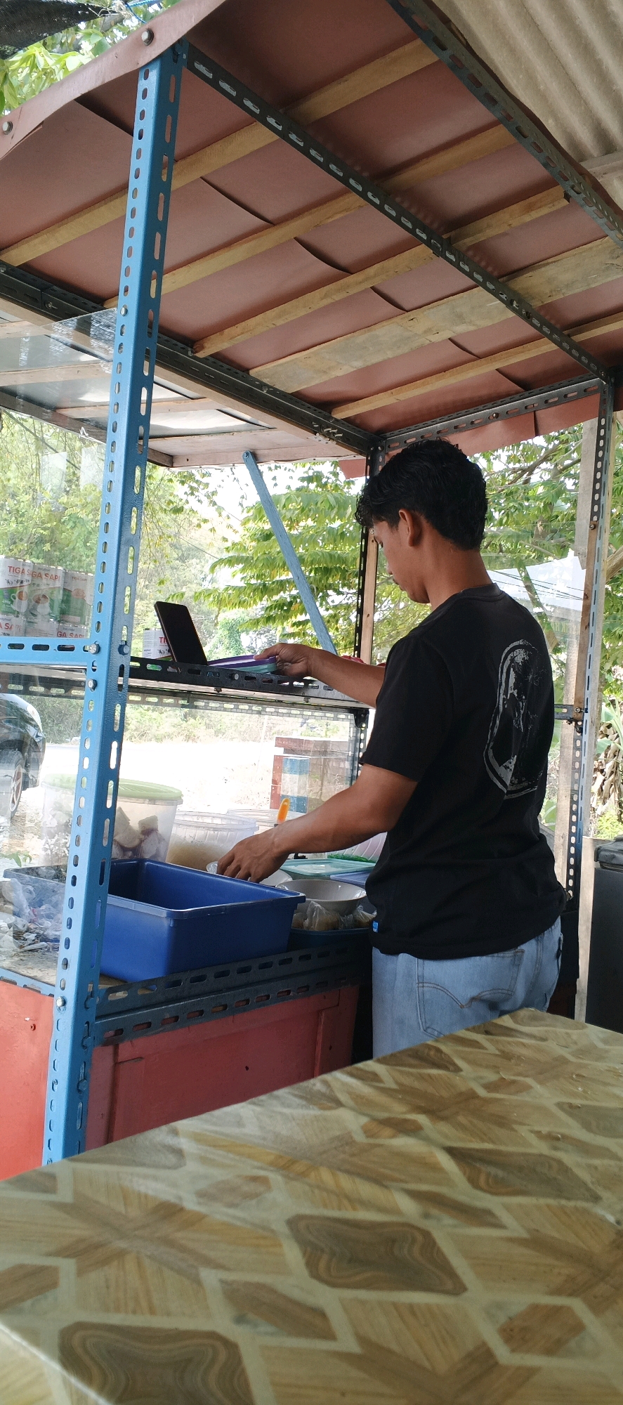 In Sonoadi, Indonesia, a young man in a black t-shirt and light blue jeans stands behind a food stall. The stall has a metal shelving unit with blue uprights and gray horizontal supports. On the shelves, there are various items, including a laptop, plastic containers, bowls, and a blue rectangular plastic tub. The man is looking down at the shelves and appears to be reaching for something or placing items. The background shows greenery and trees, indicating an outdoor setting. The foreground features a table with a patterned surface, likely a countertop or a separate table for customers. The overall impression is of a busy food stall or small eatery in a public area.