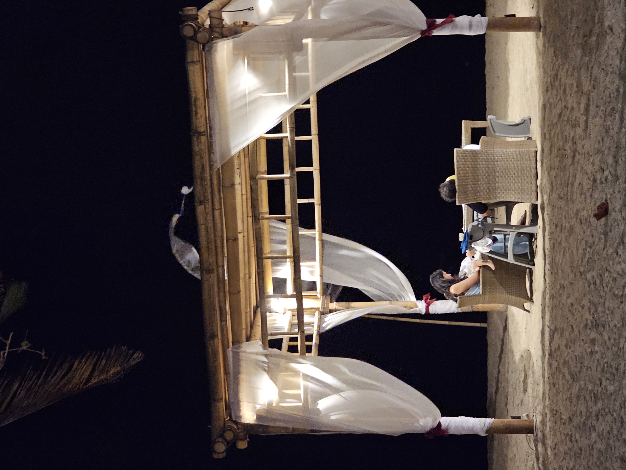 An outdoor dining structure made of bamboo and draped with white sheer fabric stands on a sandy beach at night. Two individuals are seated at a table under the structure. One person, a woman with dark hair, is sitting in a wicker chair on the left side of the table, appearing to be looking down at something on the table. Another person, with short dark hair and wearing a yellow cap, is seated in a wicker chair on the right side of the table, facing away from the camera. The environment is dark, with the only light sources appearing to be inside the dining structure, illuminating the fabric and creating shadows. The background is black, indicating the night sky. The beach sand is visible in the foreground and around the structure. The location is the city of Denpasar, Indonesia.