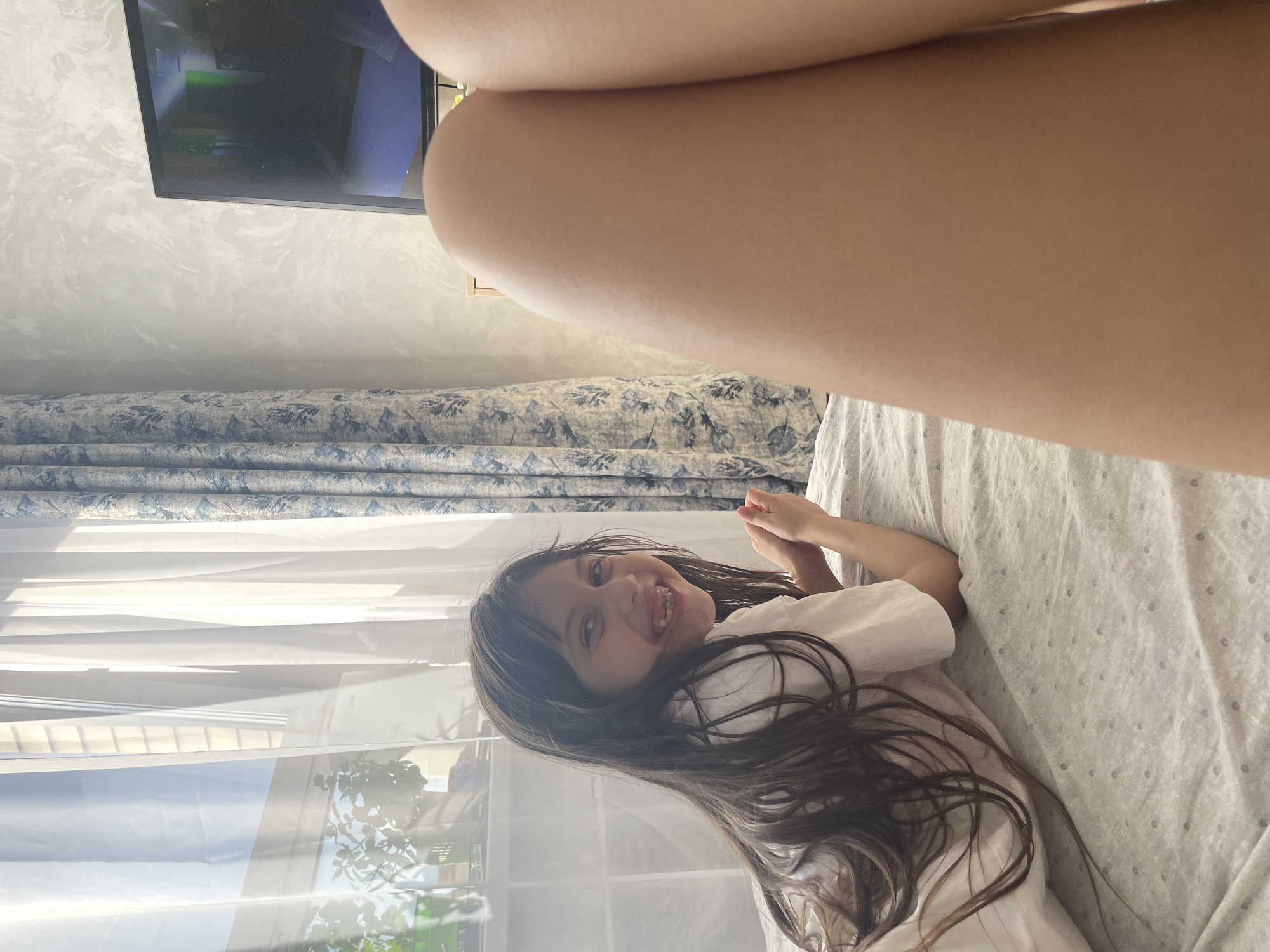 This indoor photograph captures a playful moment, likely in a bedroom in Izhevsk, Russia, during the day, indicated by bright sunlight illuminating the scene through a window.
The main subject is a young girl with long, dark hair, lying on her side on a bed. She is wearing a white t-shirt and is looking towards the camera with a wide, joyful smile, revealing her teeth. Her expression suggests happiness and playfulness. The photo is taken from a high angle, with a person's bare leg partially visible in the upper foreground, suggesting the photographer is positioned above or near the girl.
The bed is covered with a light-colored sheet featuring a subtle dotted or geometric pattern. In the background, a flat-screen television is mounted on a wall with a light grey, textured finish, possibly stucco or wallpaper with a swirling, marble-like pattern. The TV screen displays content with visible green and blue areas, potentially from a cartoon or video game. Below the television, patterned curtains, predominantly blue, white, and grey with a floral or botanical design, are drawn. A window behind the girl is partially covered by translucent white blinds or curtains, and reflections of outdoor foliage or plants are visible through the glass. No visible text is present in the image.