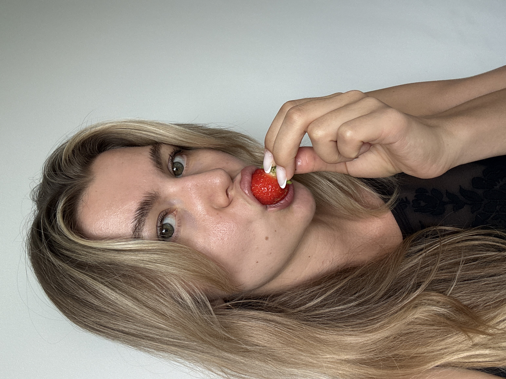 A young Caucasian woman with blonde hair is shown from the chest up against a plain white background. She has green eyes, light skin with a few freckles, and is wearing a black top with a floral lace pattern. She is holding a single red strawberry to her lips with her right hand, her mouth partially open. Her fingernails are painted with a French manicure. The lighting is soft and even.