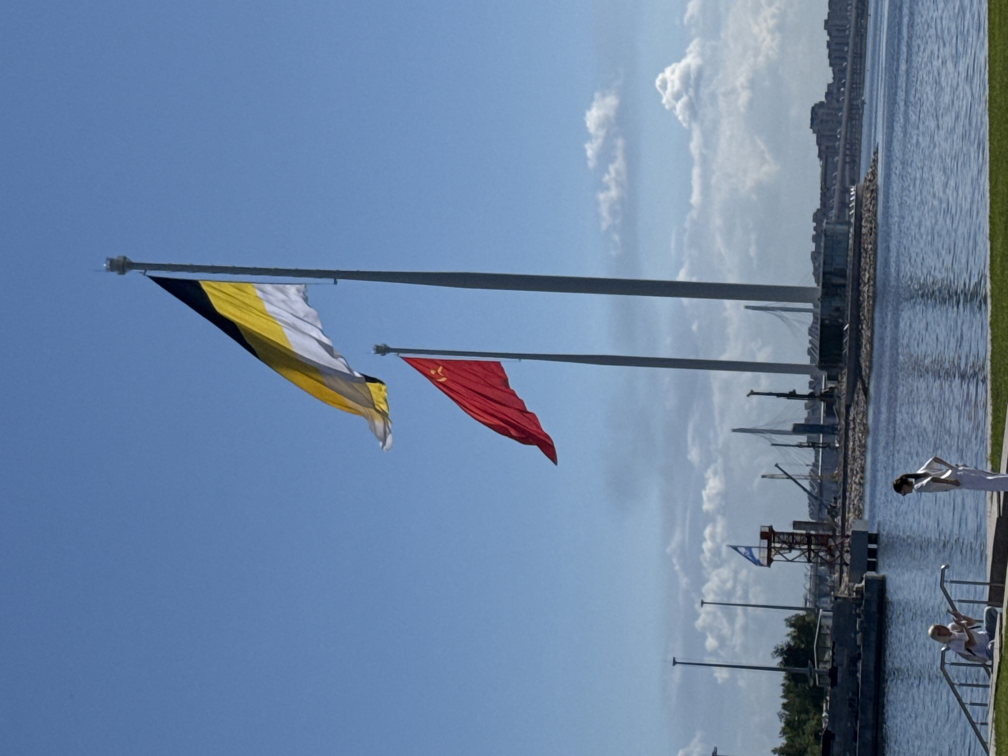 The image, taken in Saint Petersburg, Russia, presents a vibrant daytime scene dominated by a clear blue sky and a calm body of water. In the foreground, two prominent flags are flying from tall, white flagpoles. The flag on the left displays horizontal stripes of yellow, black, and white, recognized as the historical flag of the Russian Empire. To its right, the national flag of China, a red banner adorned with yellow stars, flies alongside it. Both flags are unfurled by what appears to be a gentle breeze, creating dynamic folds in the fabric.
To the right, a wide expanse of water, likely a river or bay, stretches into the distance, reflecting the bright sky. The far bank features a cityscape with numerous buildings, suggesting an urban environment. A strip of well-maintained green grass borders the water in the foreground on the right. Further in the midground, several industrial structures, possibly cranes or port facilities, are visible near the water's edge. A small flag with blue and white colors can be seen flying from a structure near these facilities.
Two individuals are positioned on the right side of the image, near the water. One person, dressed in white, stands next to a metal railing at the water's edge. Below them, another person, also in light-colored attire, is seated on a set of steps or a similar structure that descends into the water. The weather is sunny with a few scattered white clouds visible in the sky. There is no visible text in the image.