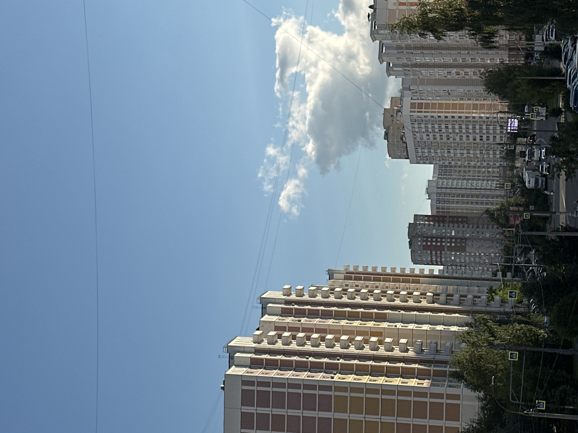 A wide shot captures a street scene in Podolsk, Russia, under a clear blue sky with scattered white clouds. The foreground is dominated by a dense canopy of lush green trees on the left and right sides of the frame. A road curves through the center, with several vehicles, including a white truck, a white car, and other vehicles, visible. Pedestrians can be seen walking on the sidewalk. Yellow pedestrian crossing signs are mounted on poles along the road. In the background, a row of tall, multi-story apartment buildings with repetitive window patterns and balconies stretches across the horizon. Power lines crisscross the sky above the buildings.