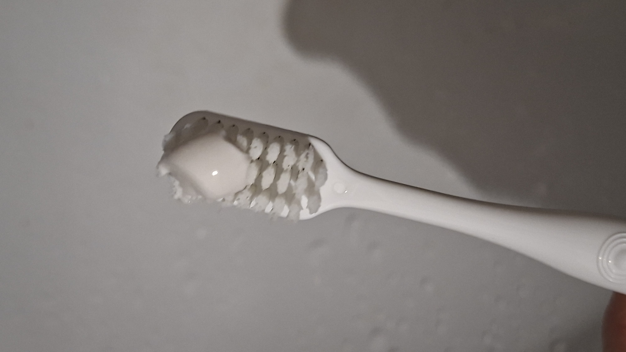 A close-up, eye-level shot shows a white toothbrush with a blob of white toothpaste on its bristles. The toothbrush is positioned vertically, with its handle extending downwards and out of frame at the bottom left. The bristles are white and appear slightly worn, with some dark specks visible among them. The toothpaste is a thick, creamy substance, forming a rounded dollop on the bristles. The background is a neutral, textured gray wall, with a soft shadow of the toothbrush cast to the right. There are no people or other objects visible in the scene. The setting appears to be a bathroom or similar environment. There is no indication of the location being in Забалоцкі сельскі Савет, Belarus.