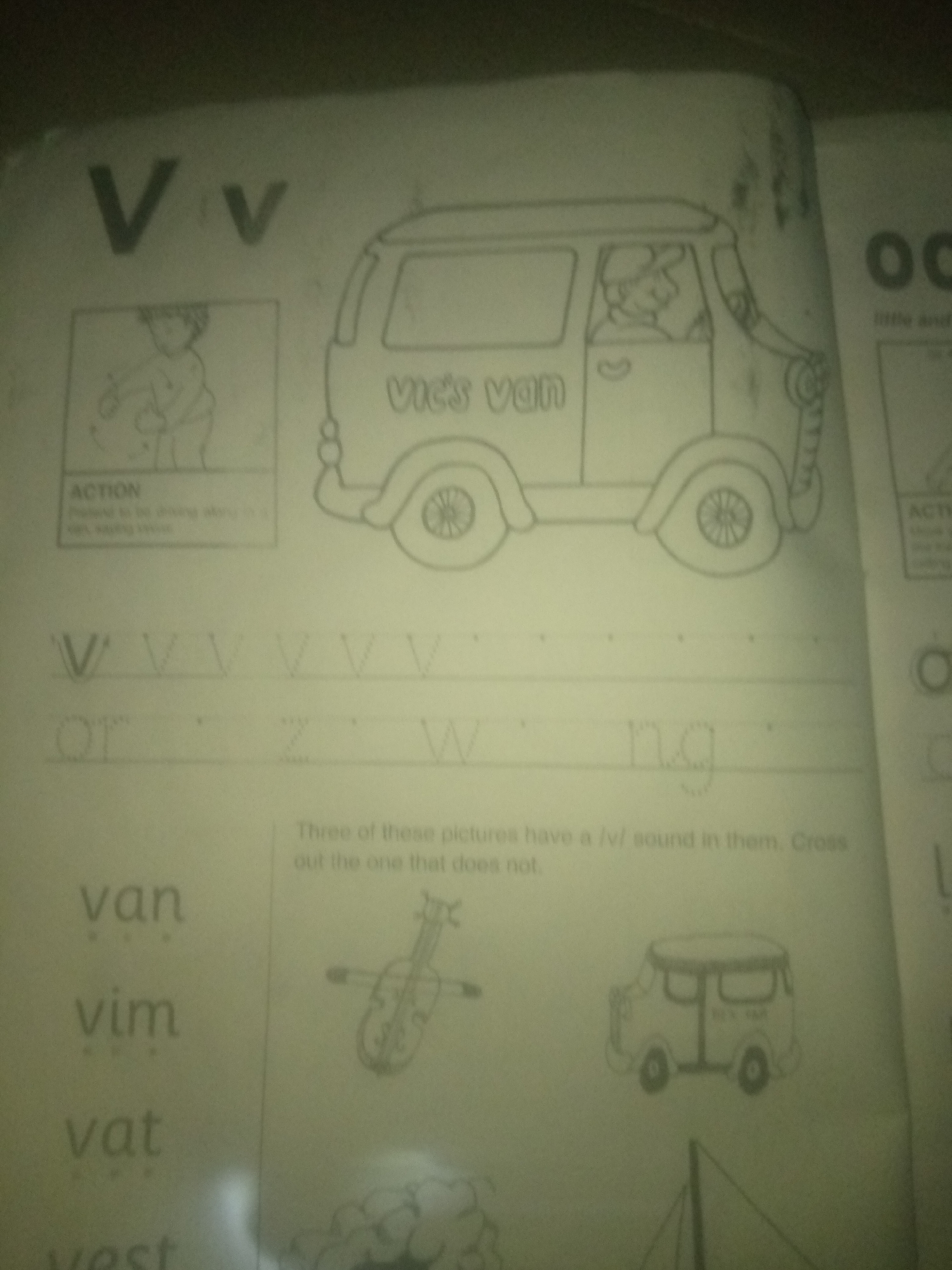 The image shows a page from a children's alphabet and reading practice book. The page features the letter "V" prominently at the top. Below "V" is a drawing of a van with the words "Vic's van" written on its side. To the left of the van is a small drawing of a person appearing to be in motion, with the word "ACTION" below it. Further down the page, there are dotted lines for tracing the letter "V" multiple times. Below this, there are the words "van", "vim", and "vat" written, each with dotted lines beneath them. To the right of these words are drawings of a violin and another van. A sentence above these images reads, "Three of these pictures have a /v/ sound in them. Cross out the one that does not." There is also a faint drawing of a boat at the bottom right of the page. The image is taken from an angle and appears to be a photograph of a physical page, not a digital illustration. There are no indications of the city of Jauro Yinu, Nigeria, or any specific urban environment.