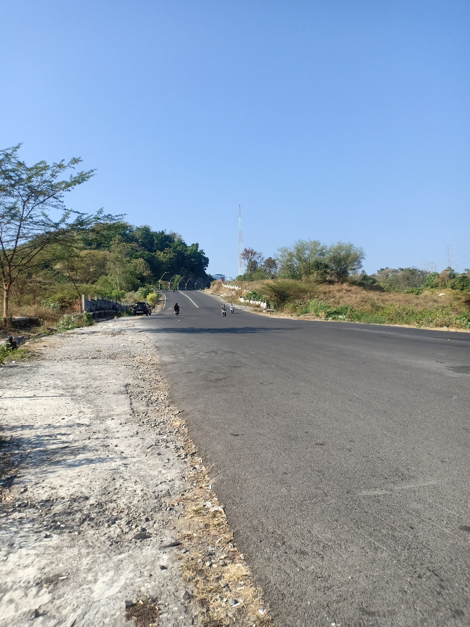 The image shows a road ascending a gentle incline in a rural or semi-rural area. The road surface is asphalt and appears to be well-maintained. On the left side of the road, there is a rough, unpaved shoulder with some debris and dry vegetation. Further up the incline, there are some trees and lush greenery on a hillside. A tall, red and white telecommunications tower is visible in the distance on the left. On the right side of the road, the landscape is characterized by dry grass and scattered trees. There are a few individuals on motorcycles traveling up the road, and a dark-colored car is parked on the side of the road. The sky is clear and blue, suggesting a sunny day. This scene is consistent with a location on the outskirts of Semarang, Indonesia, possibly along a route leading into the hills.