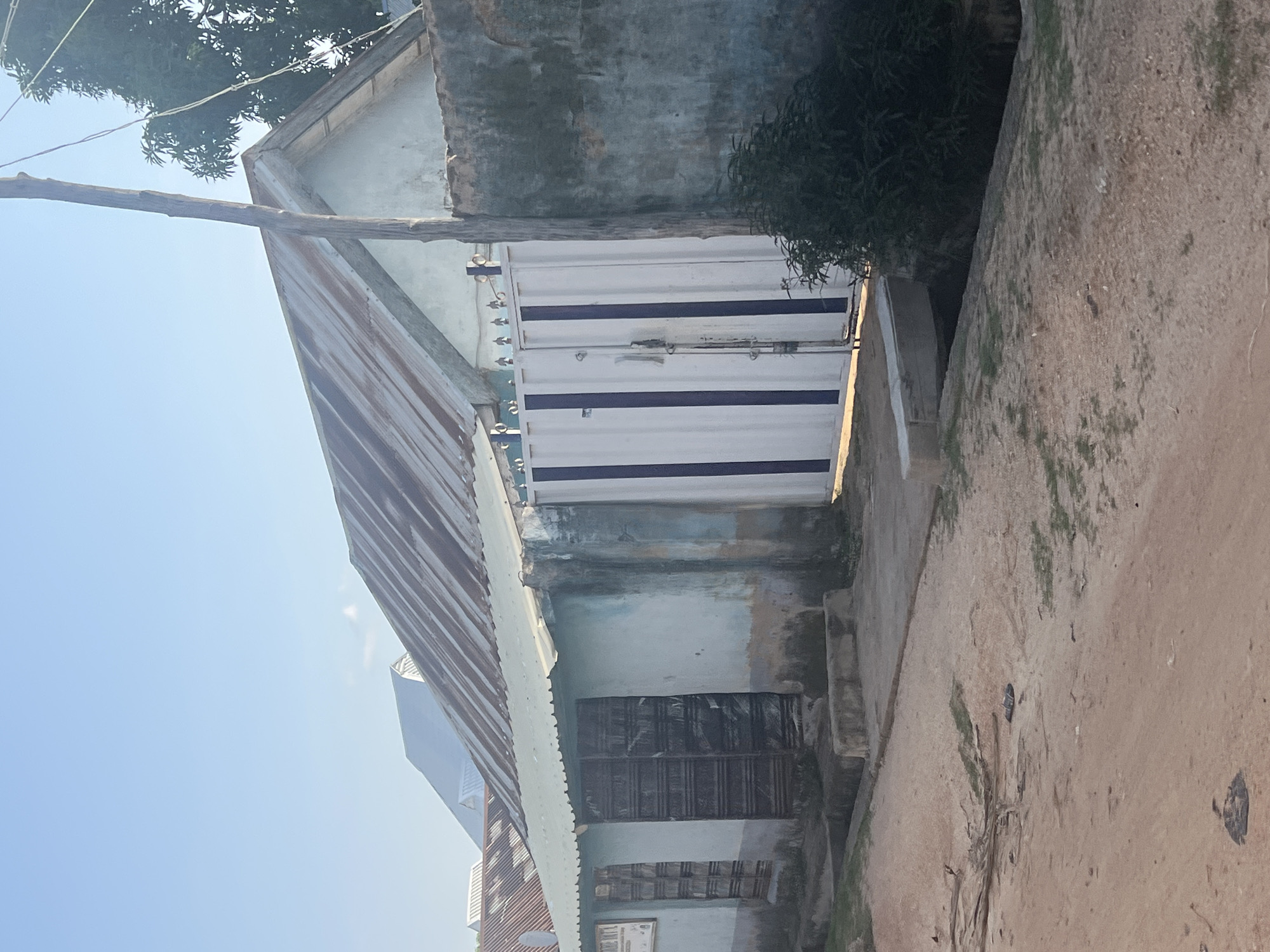 The image depicts a street scene in Jalingo, Nigeria. The foreground shows a dirt road with scattered debris and a pair of sandals. In the mid-ground, a light blue building with a corrugated metal roof stands. The building has two large, dark metal doors and a white gate with blue vertical stripes. Adjacent to the building is a rough, dark-colored wall. A wooden pole stands in front of the wall. Behind the building, other structures with corrugated metal roofs are visible. The sky is clear and blue.