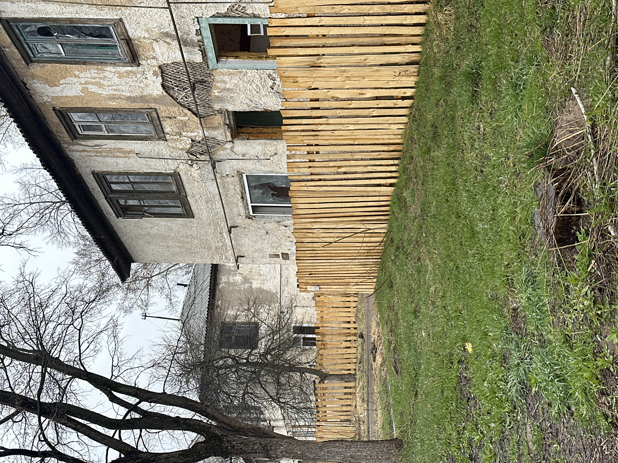 The image, rotated 90 degrees clockwise, depicts a dilapidated, two-story building on the left, an expanse of grass on the right, and a newly constructed wooden fence running between them.
The old building features a textured, peeling facade with visible damage, exposing underlying lath or brickwork. Multiple windows are present; some are broken with shattered panes, while others appear intact but dirty. A small opening or vent is visible on the upper floor. The roofline of a lower section of the building, possibly an extension, is visible with what appears to be dark corrugated metal.
A light-colored wooden fence, made of horizontal planks, runs parallel to the building, separating it from the grassy area. Its fresh appearance suggests recent construction.
To the right of the fence, the ground is covered with sparse green grass mixed with patches of exposed dark earth and dry, brown vegetation. A natural depression or ditch is visible on the far right.
In the foreground and mid-ground, on the left side of the frame, several large, bare deciduous trees with gnarled branches are prominent against an overcast, light-grey sky, indicating late winter or early spring. Another smaller, bare tree stands closer to the building in the background.
The scene is static, with no human presence or activity. The overcast sky suggests daytime. The overall atmosphere conveys neglect and the passage of time, juxtaposed with the new fence. No readable text is visible on any objects in the image. The setting is consistent with a rural or semi-urban environment in Chaikovsky, Russia.