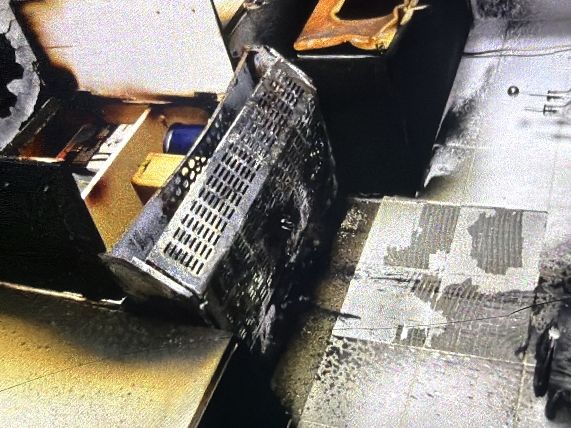 The image depicts the aftermath of a fire in what appears to be a kitchen or storage area, possibly in Bukuru, Nigeria. A metal grate basket, likely from a ventilation system or a storage rack, is heavily damaged and covered in soot and debris. It is suspended at an angle, showing signs of melting and deformation. Below it, the wall tiles are discolored and some are peeling away, indicating exposure to intense heat. Dark soot stains are visible on the walls and ceiling, with some areas appearing charred. In the background, shelves are visible, holding what appear to be boxes and a blue container, also affected by the fire. The overall scene conveys significant fire damage.