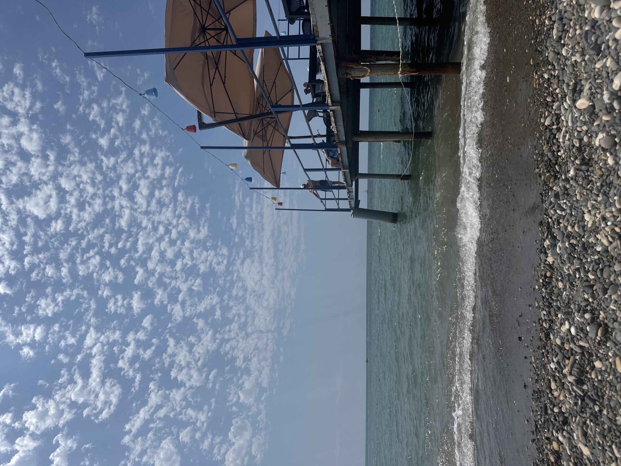 The image depicts a beach scene with a pier extending over the water. The foreground is a pebbled beach with small waves gently washing ashore. The sea is a deep green, and the horizon is clear, with a small boat visible in the distance.
The pier is constructed with wooden planks and supported by metal and wooden pilings. Several large, tan umbrellas provide shade over tables and chairs on the pier. A string of colorful decorative lights, shaped like bells, hangs above the pier.
On the pier, a man stands by the railing, facing away from the camera and towards the sea. Further along the pier, several other people are seated at tables, appearing to be patrons of an establishment on the pier. The sky is a bright blue, filled with scattered, fluffy white clouds. The overall impression is of a sunny day at a seaside resort or cafe. The location is identified as Sokhumi, Abkhazia.