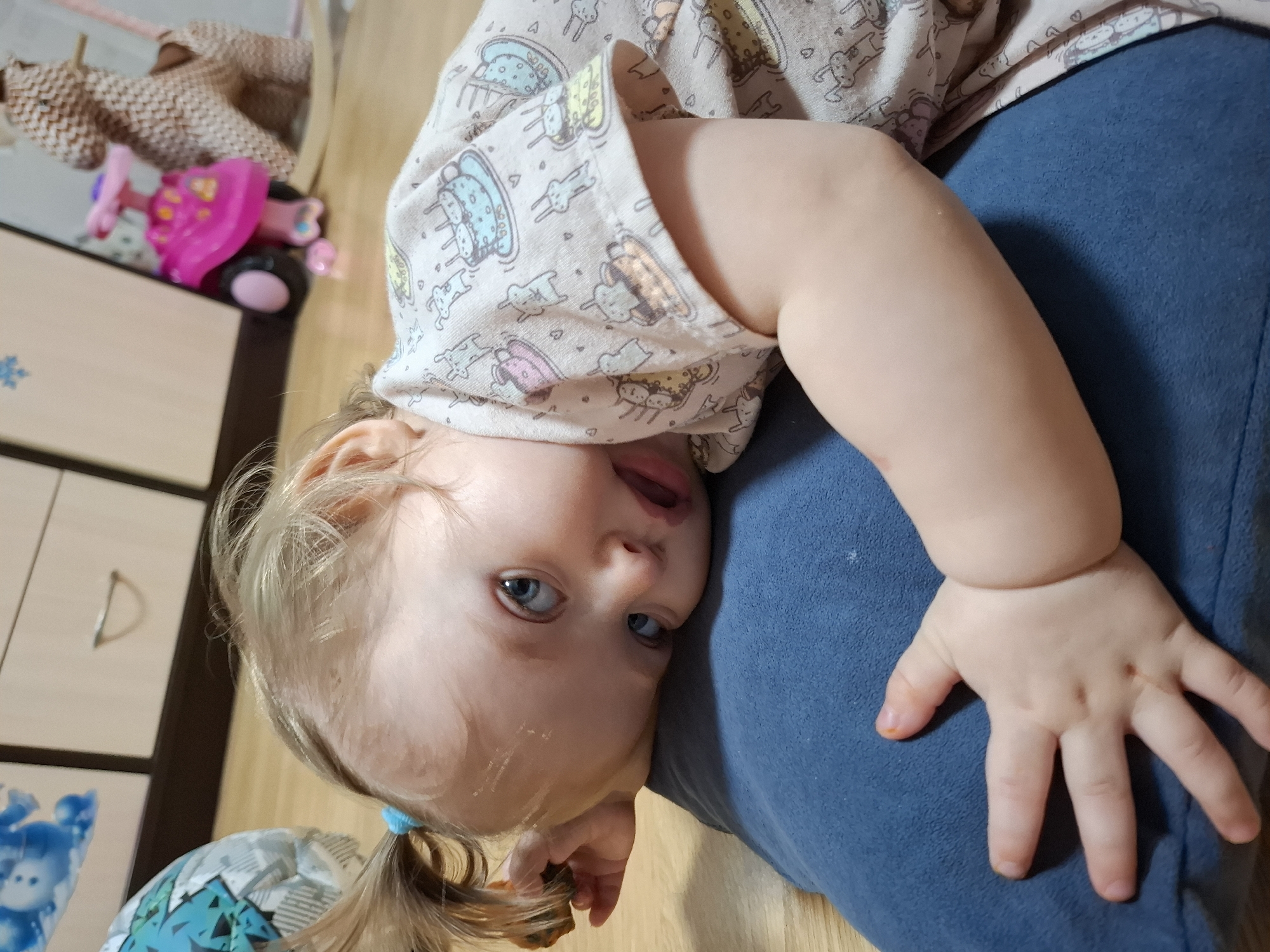 A young child with fair skin and blue eyes is lying on a blue surface, possibly a pillow or cushion. She is wearing a light-colored t-shirt printed with cartoon designs. The child's hair is blonde and gathered in a small ponytail.
In the background, there's a wooden floor and a dresser. A toy tricycle and a stuffed animal are visible on the top of the dresser.
Given the city specified, this image was likely taken in Chaikovsky, Russia. There is no information to suggest the precise location of the image.