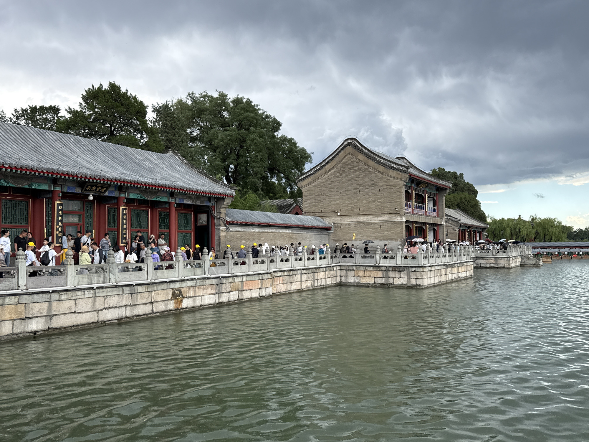 This image depicts a section of the Summer Palace (Yiheyuan) in the Haidian District of Beijing, China, specifically along the shore of Kunming Lake.
In the foreground, a large body of water, likely Kunming Lake, exhibits numerous ripples across its dark green surface, reflecting the overcast sky. A stone promenade with an ornate grey railing runs horizontally across the midground. The railing features carved panels and is supported by a robust retaining wall composed of large, light-colored rectangular stone blocks at the water's edge.
Approximately 40 to 50 individuals are visible on the promenade, walking in both directions. Many are wearing casual clothing, and several in the left portion of the promenade are distinguishable by bright yellow hats. Some individuals are holding open umbrellas, suggesting either sun protection or a recent/imminent light rain.
Lining the promenade are several traditional Chinese buildings. On the left, a long, single-story building with a grey tiled, ridged roof is prominent. Its facade is characterized by multiple red columns, dark wooden window frames, and decorative elements under the eaves. A horizontal plaque above an entrance on this building displays Chinese characters, which are identifiable as "景福閣" (Jingfu Ge, or Pavilion of Scenery and Blessing). Adjacent to it on the right is a taller, two-story structure constructed from grey bricks, featuring a distinctive curved grey tiled roof. This building also presents balconies with red balustrades on its upper level. Further to the right, similar traditional buildings extend along the waterfront.
Behind the structures, dense green trees are visible, and on the far right, beyond the buildings and across the water, parts of a bridge structure and additional traditional architecture are discernible amidst more foliage. The sky above is heavily overcast with dark grey clouds, though a patch of lighter blue sky is visible on the far right horizon.