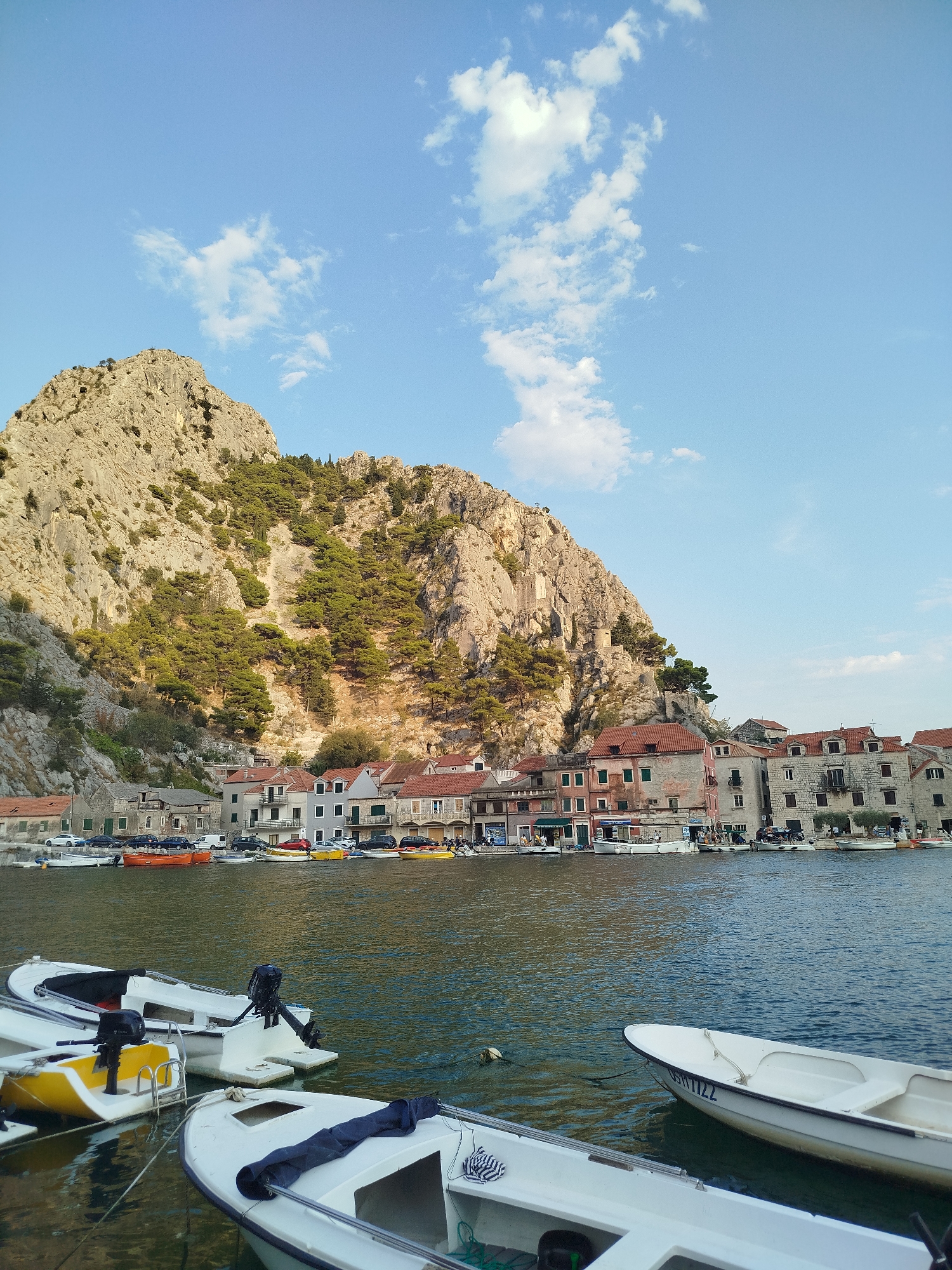 A wide, long shot captures a serene waterfront scene in Omiš, Croatia. The foreground features several small white boats docked together, their motors visible and one with a blue cloth draped inside. The water, a deep blue-green, gently ripples, reflecting the sky and the buildings. Beyond the boats, a row of charming, old stone buildings with terracotta roofs lines the waterfront. These buildings appear to be shops or residences, with awnings and windows suggesting activity within. Cars are parked along the street in front of the buildings. Dominating the background is a dramatic, sheer limestone cliff face, sparsely covered with green pine trees. The sky above is a clear blue, punctuated by scattered white clouds. The lighting suggests it is late afternoon or early evening, with the sun casting a warm glow on the scene.