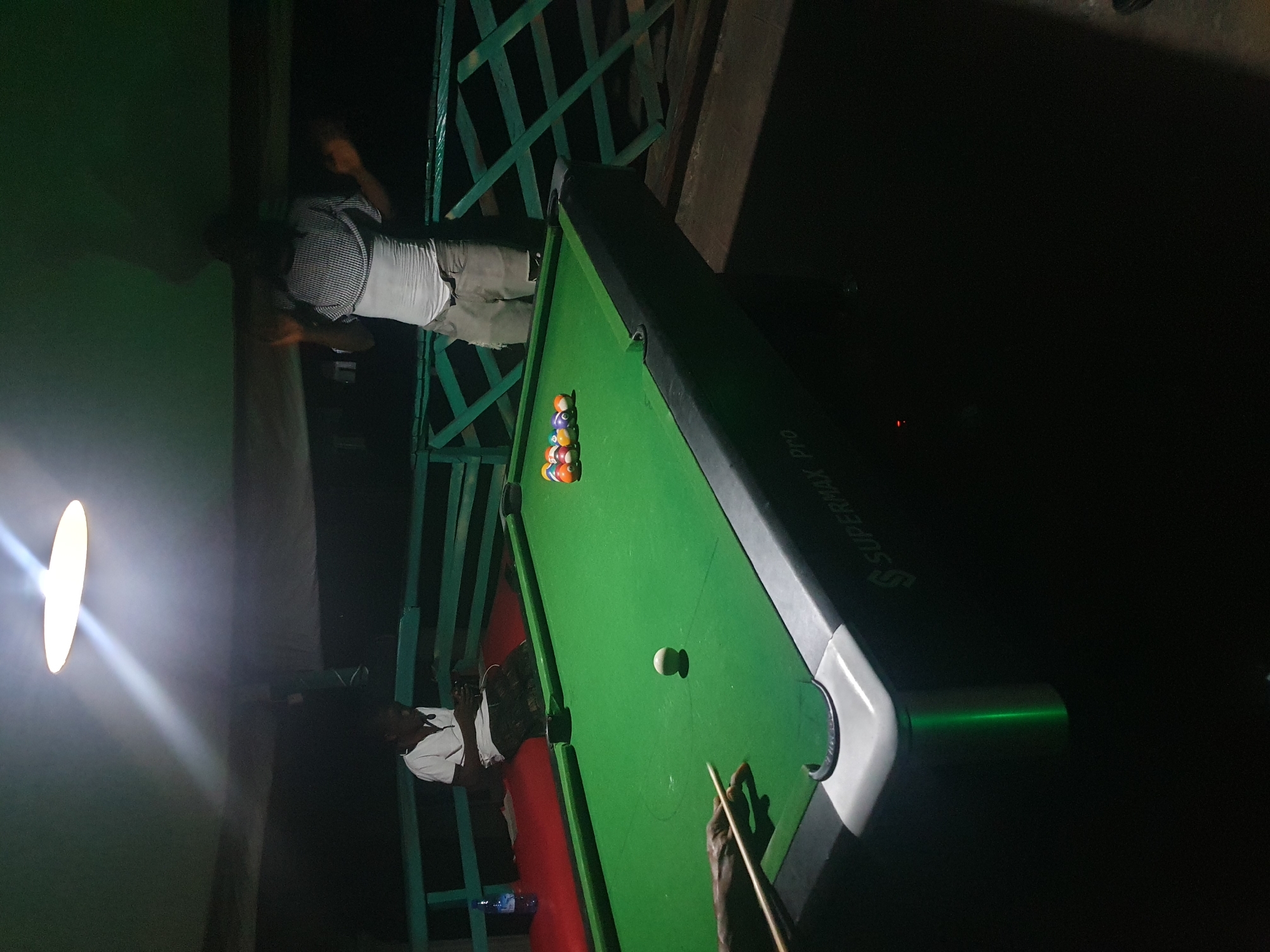 In Lokoja, Nigeria, two men are present in a dimly lit outdoor setting, likely a bar or recreational area, around a billiard table. One man, wearing a white t-shirt and patterned trousers, is seated on a red surface, looking down at a mobile device. The other man, dressed in a white tank top and grey distressed shorts, stands near the billiard table, holding his hands up, possibly in a gesture of surprise or excitement. A rack of billiard balls, including a white cue ball, is arranged on the green felt surface of the table. The table itself has a "SUPERMAX Pro" marking. The environment features a green railing and a bright overhead light.