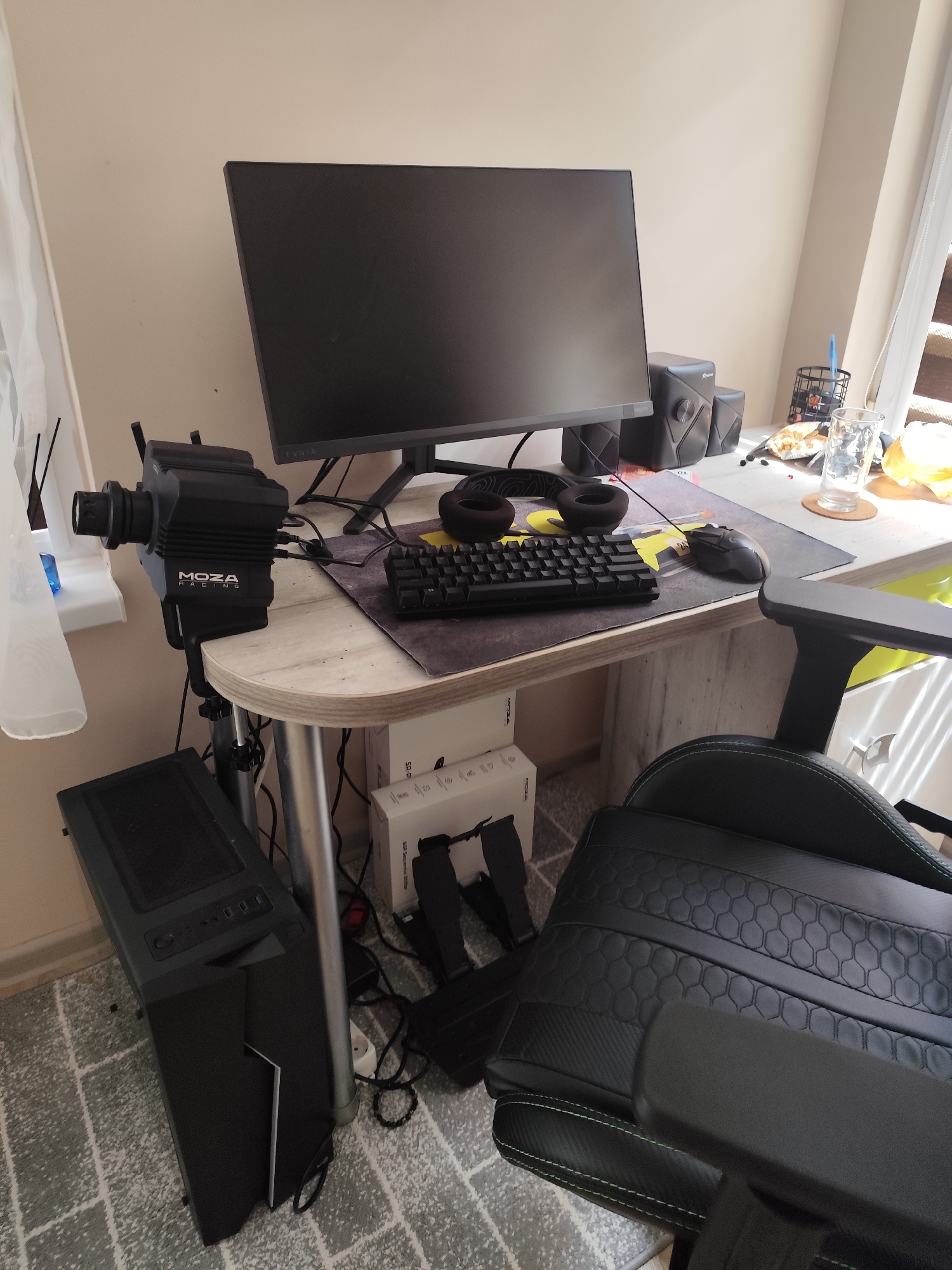 The image depicts a home office or gaming setup. A black computer tower is positioned on the floor to the left of a light-colored, curved desk. On the desk, a large black computer monitor is centrally placed. To the left of the monitor, a black racing wheel controller is clamped to the desk. Headphones are placed on the desk in front of the monitor, next to a black keyboard. A computer mouse is to the right of the keyboard. Two black speakers are situated behind the monitor. A glass of water and a pen holder with pens are visible on the right side of the desk. A black gaming chair with green accents is in the foreground, partially obscuring the desk. The room has a light-colored wall and a window with sheer curtains on the left. The floor is covered with a grey and white patterned rug. The scene suggests a dedicated space for PC gaming or sim racing. There is no information in the image to suggest the location is in the city of Pomorie, Bulgaria.