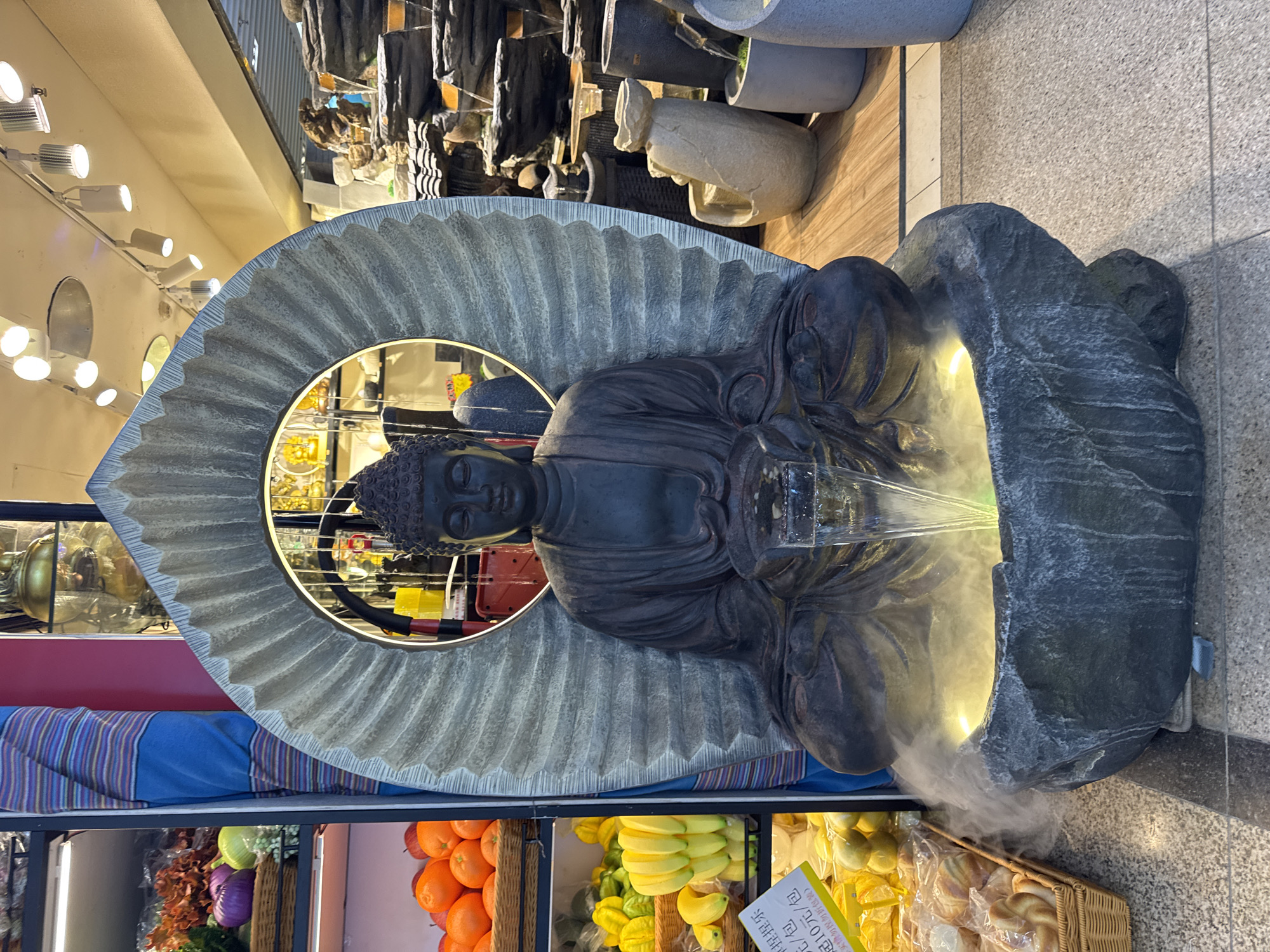 The image shows a Buddha statue fountain in an indoor market setting, possibly in the Yuexiu District of China. The statue is a dark gray color, positioned against a light gray, fan-shaped backdrop. Water is flowing from the statue's hands into a basin, creating a mist-like effect around the base. Below the fountain, there are shelves displaying various food items, including fruits like oranges and bananas, along with baked goods in a basket. The background reveals additional decorative items, including stacked pots and other statues, as well as overhead lighting fixtures. The environment suggests a commercial space with a mix of decorative and functional elements.