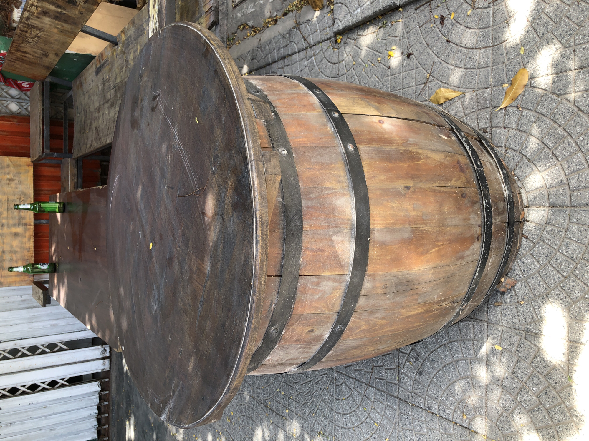 This image depicts an outdoor setting in Đà Nẵng, Vietnam. The primary subjects are a large, round wooden table with a dark finish, and a barrel-shaped structure that appears to be supporting one side of the table, or possibly serving as a decorative element. The table is situated on a paved area with a distinctive circular pattern, interspersed with fallen leaves.
Two green glass bottles, likely beer bottles, are placed on the table, suggesting a casual or social environment, perhaps a cafe or outdoor dining area. The table itself has a worn appearance, with visible scratches and a sheen indicating it has been used. The barrel is constructed from wooden staves and reinforced with dark metal bands.
The scene is illuminated by natural sunlight, casting shadows on the ground and creating a dappled effect, indicating it is daytime. The weather appears to be clear, given the strong light. The paving stones and the type of vegetation visible in the background hint at a tropical or subtropical climate, consistent with Đà Nẵng.
There is no discernible text visible in the image. The overall impression is one of an informal, possibly rustic, outdoor gathering space. The arrangement of the table and barrel suggests an unconventional design for a seating or serving area.