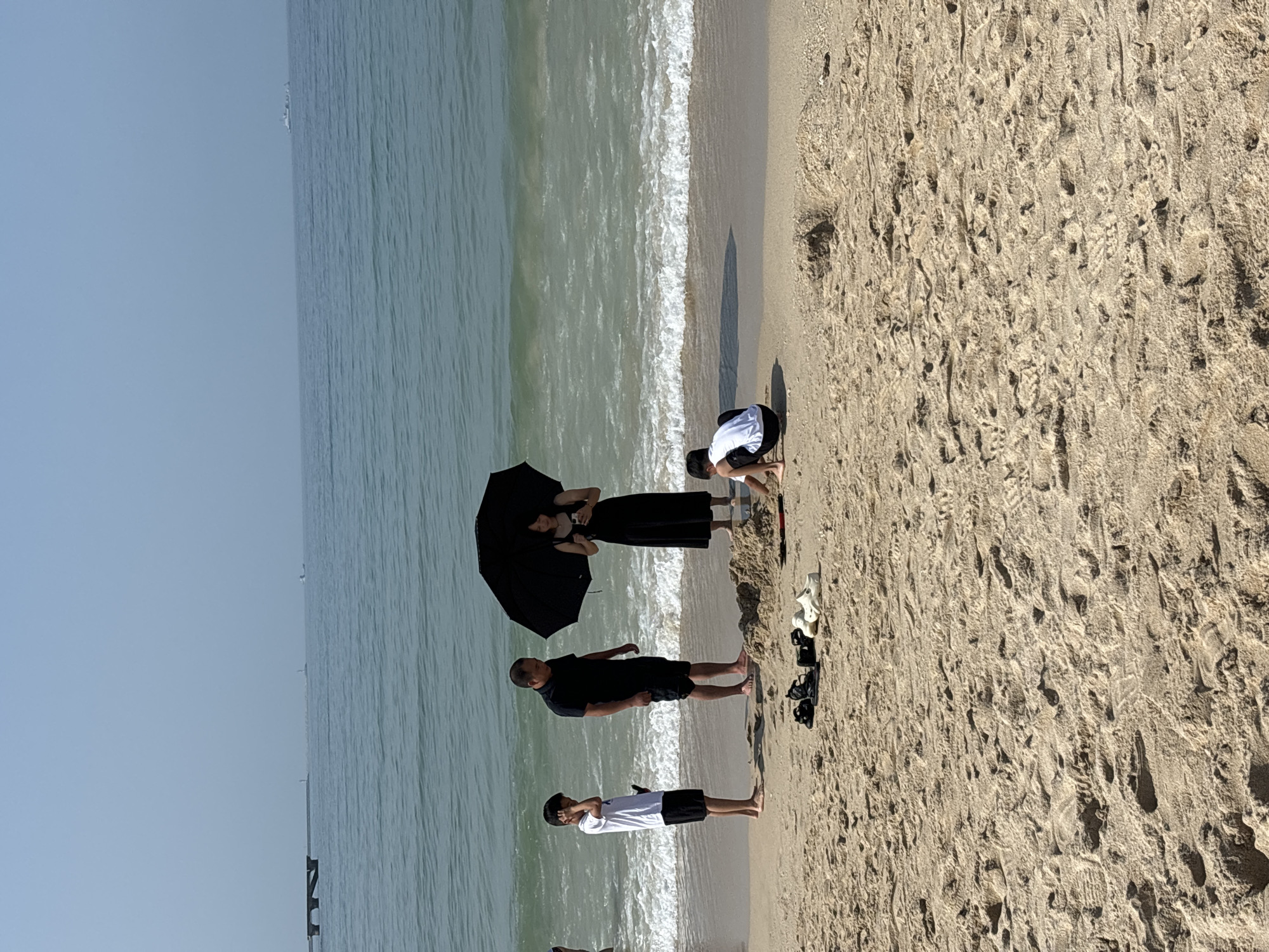 The image depicts a beach scene in Jiyang District, China, with four individuals present. A young boy in a white t-shirt and black shorts stands on the left, holding a phone. Beside him, a man in a black t-shirt and black shorts stands barefoot. A woman in a long black dress holds a black umbrella and a smartphone. In the foreground, a young boy in a white t-shirt and black shorts is crouching and playing in the sand. Several pairs of footwear, including black sandals and white slip-on shoes, are placed on the sand near the standing figures. The ocean with moderate waves is visible in the background, and a pier with a lighthouse structure extends into the water on the horizon. The sky is clear and hazy.