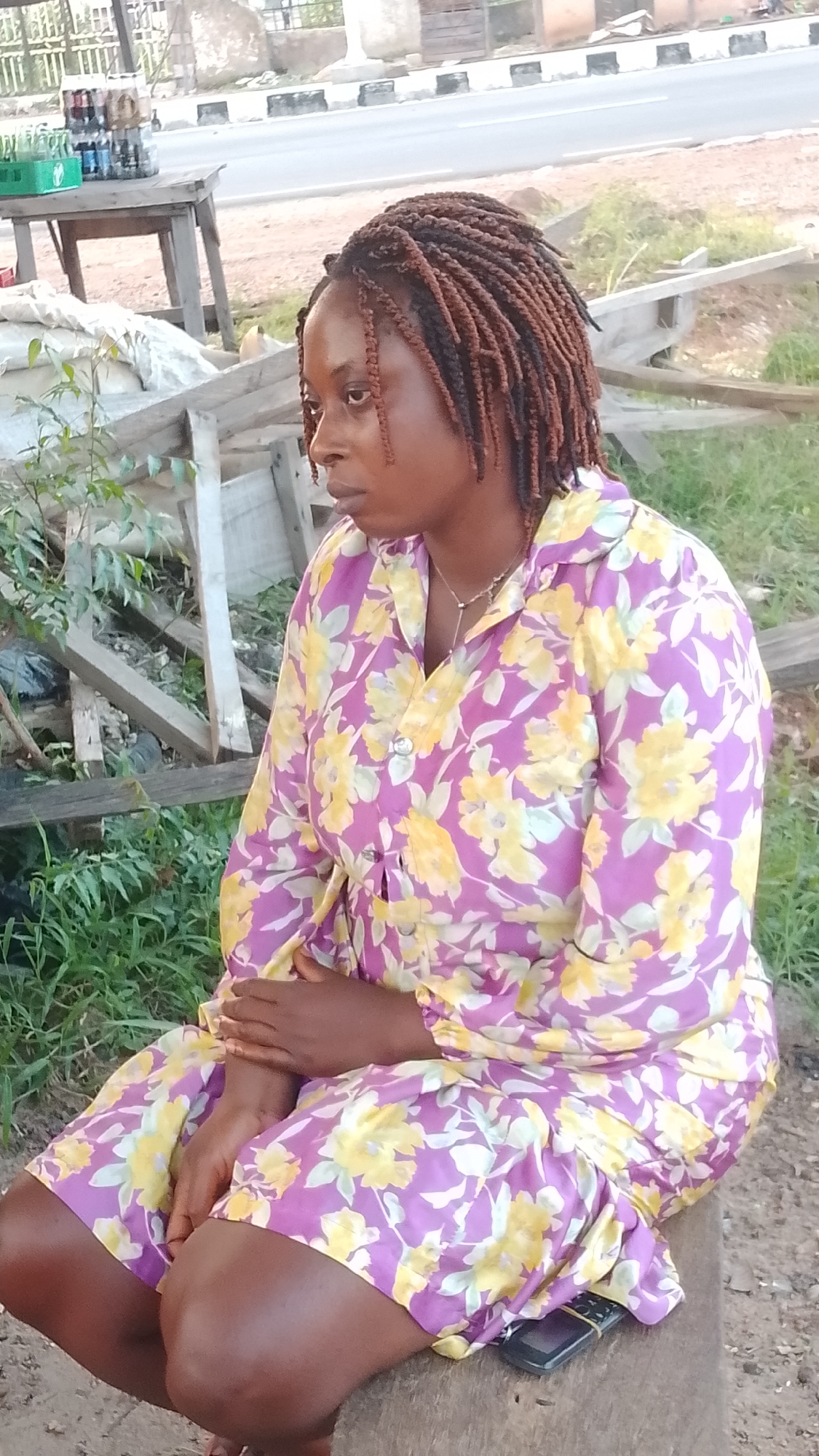 A woman with braided hair, wearing a purple and yellow floral dress, sits on a wooden bench outdoors. She is looking to her left. A mobile phone is visible under the hem of her dress. In the background, a wooden table holds several bottles of drinks, and a road is visible behind it. The scene appears to be in Adankolo, Nigeria.