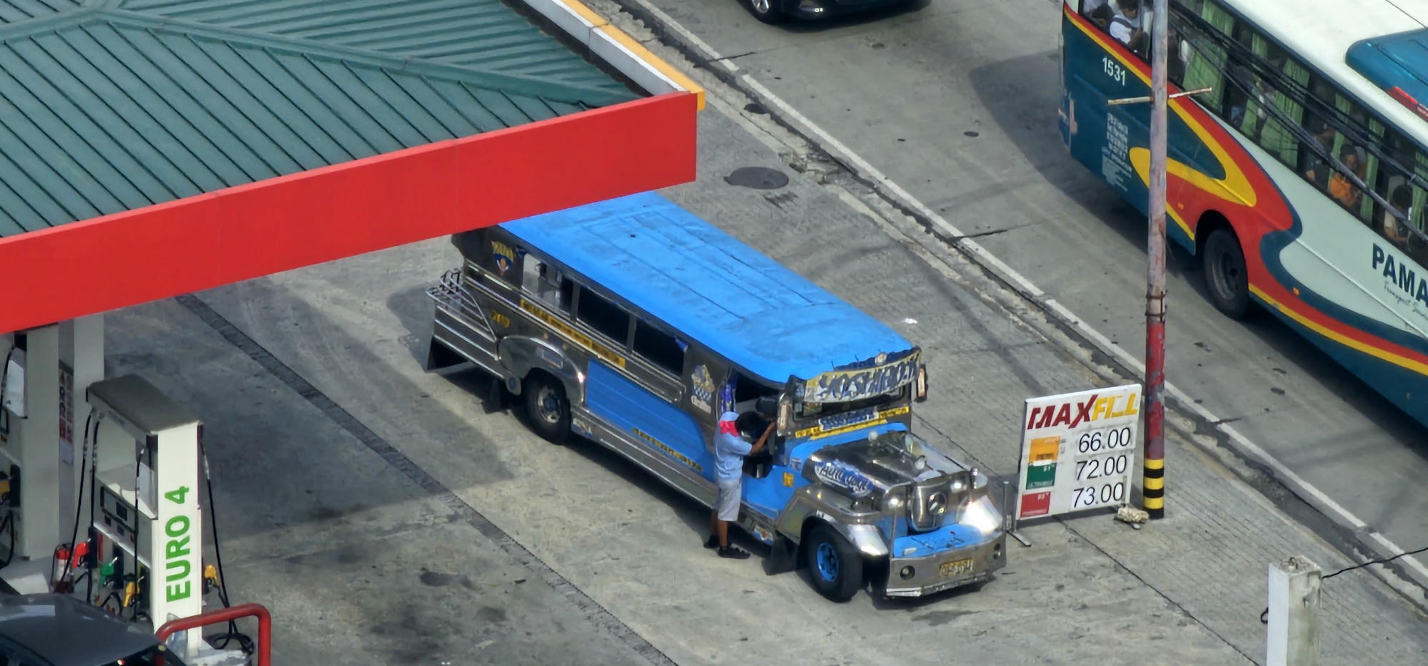 An aerial view captures a gas station in Makati, Philippines, with a bright blue jeepney and a bus as the main subjects. The jeepney, a common mode of transportation in the Philippines, is parked near a fuel pump, and a person in a light blue shirt and white shorts is seen interacting with it. The jeepney is heavily decorated with chrome and blue accents, and its roof is a vibrant blue.
To the right of the jeepney, a white bus with colorful red, yellow, and blue stripes is visible. The bus is labeled "PAMANA Transport Services." A street sign displaying fuel prices – "MAXFILL" with prices 66.00, 72.00, and 73.00 – is also present. The gas station has a green roof and a prominent red overhang. The ground is paved with concrete, and there are visible markings on the road. In the background, another dark-colored car can be seen. The scene suggests a typical day at a busy urban gas station.