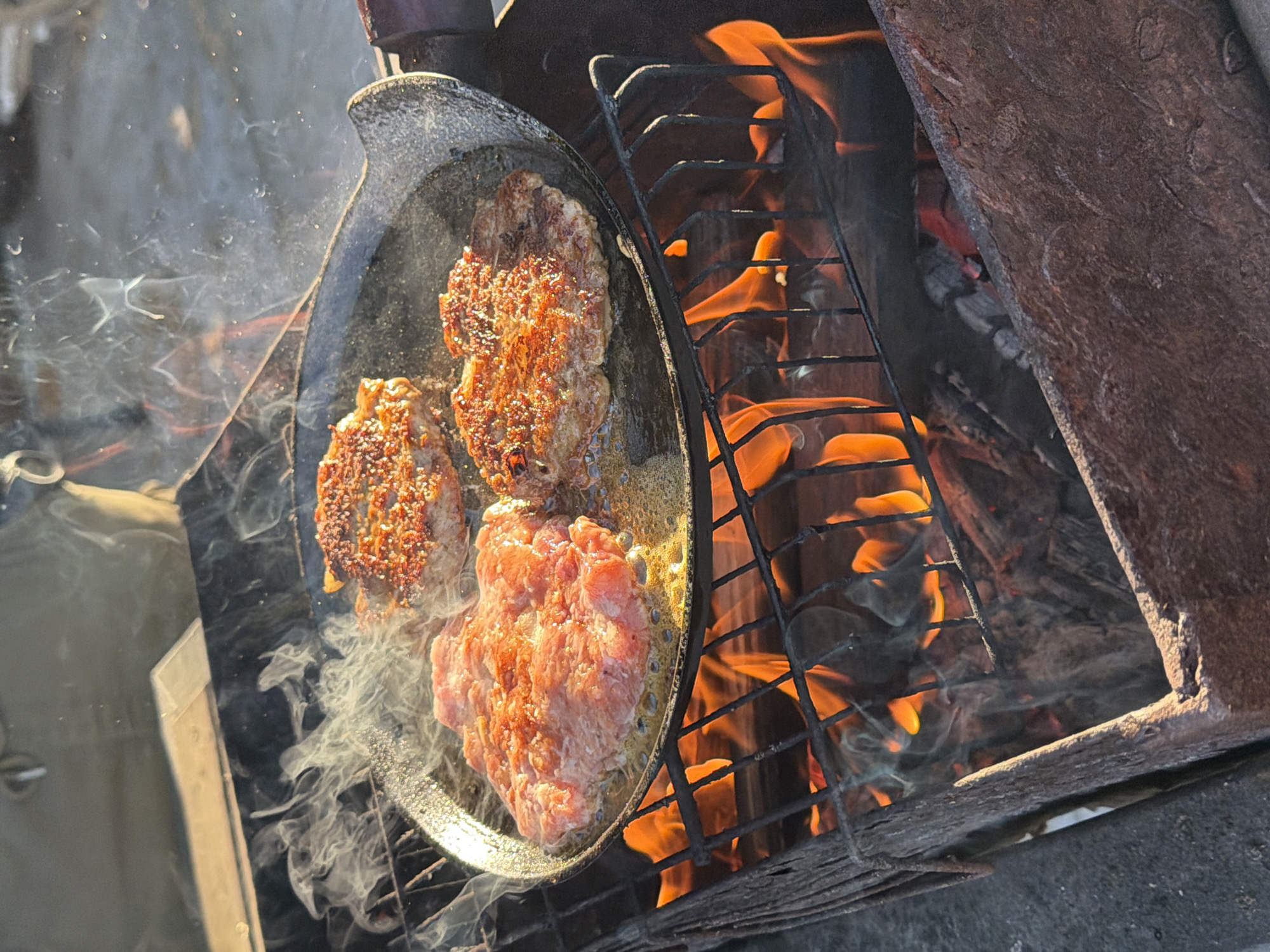 The image depicts three meat patties cooking in a frying pan over an open fire. The patties are in various stages of cooking, with one appearing raw, one partially cooked, and one cooked. Smoke is emanating from the pan. The fire is contained within a rustic metal structure. The location is Большая Шильна, Russia.