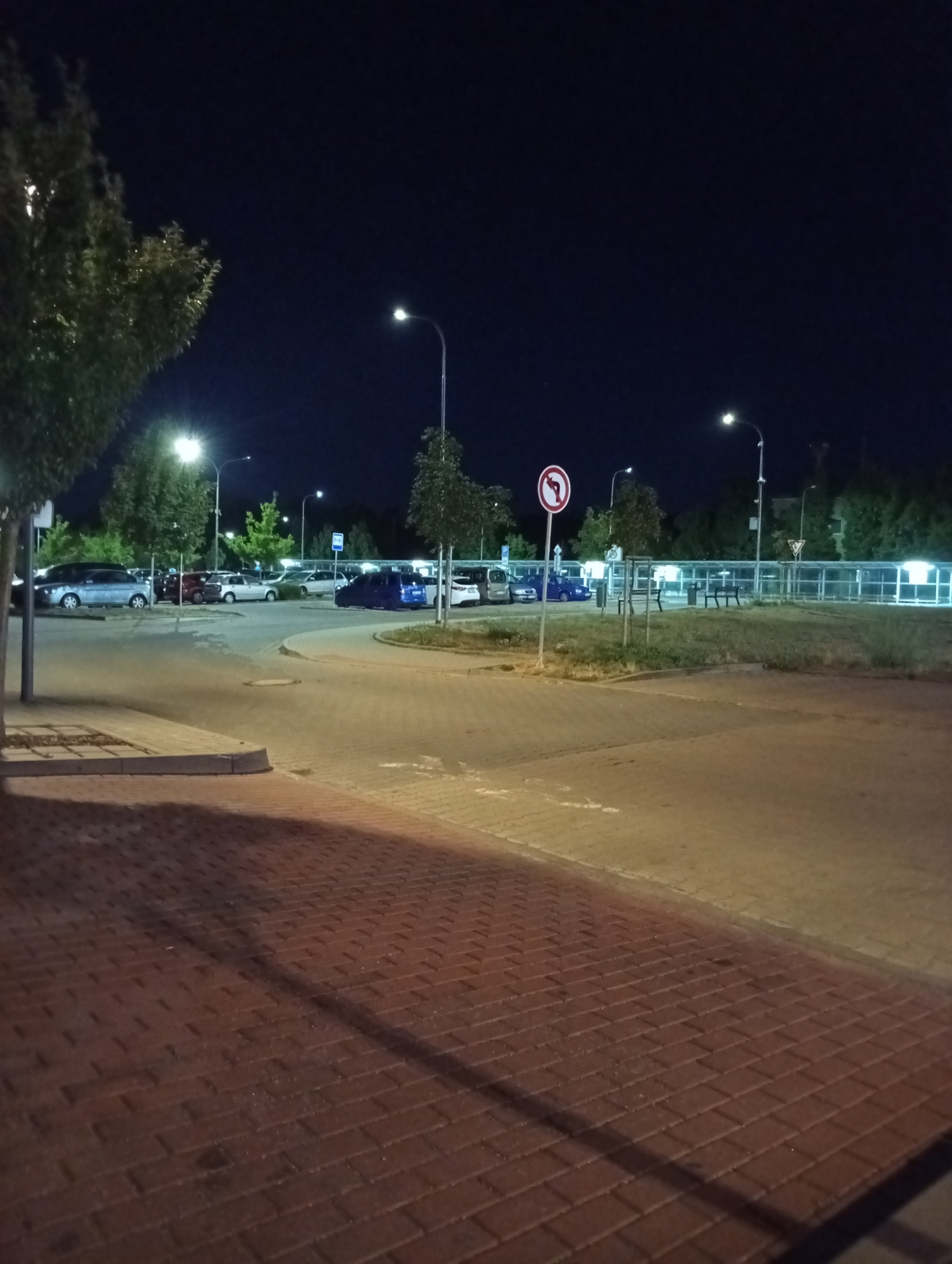 The image depicts a parking lot at night in the city of Tišnov, Czechia. The scene is illuminated by streetlights, casting long shadows on the brick pavement in the foreground. Several cars are parked in rows in the background, including a blue van. A traffic sign indicating a left turn is visible, along with a yield sign further down the road. Trees with foliage are scattered throughout the area, and a glass-enclosed structure with benches and trash cans is situated behind the parking lot. The sky is dark and clear.