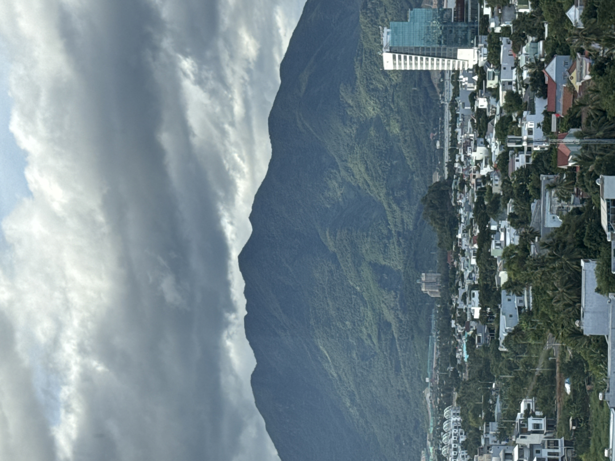 The image shows a panoramic view of a city with a large mountain range in the background, likely captured from an elevated vantage point in Đà Nẵng, Vietnam. The sky is overcast with dark, dramatic clouds. In the middle ground, a dense urban landscape is visible, featuring a mix of low-rise buildings, some with red tiled roofs, interspersed with lush green trees, including palm trees. A prominent, modern glass skyscraper stands out on the right side of the frame, contrasting with the surrounding architecture. Further in the background, the verdant slopes of the mountains dominate the scene, appearing lush and possibly forested. The overall impression is one of a city nestled between natural grandeur and urban development.