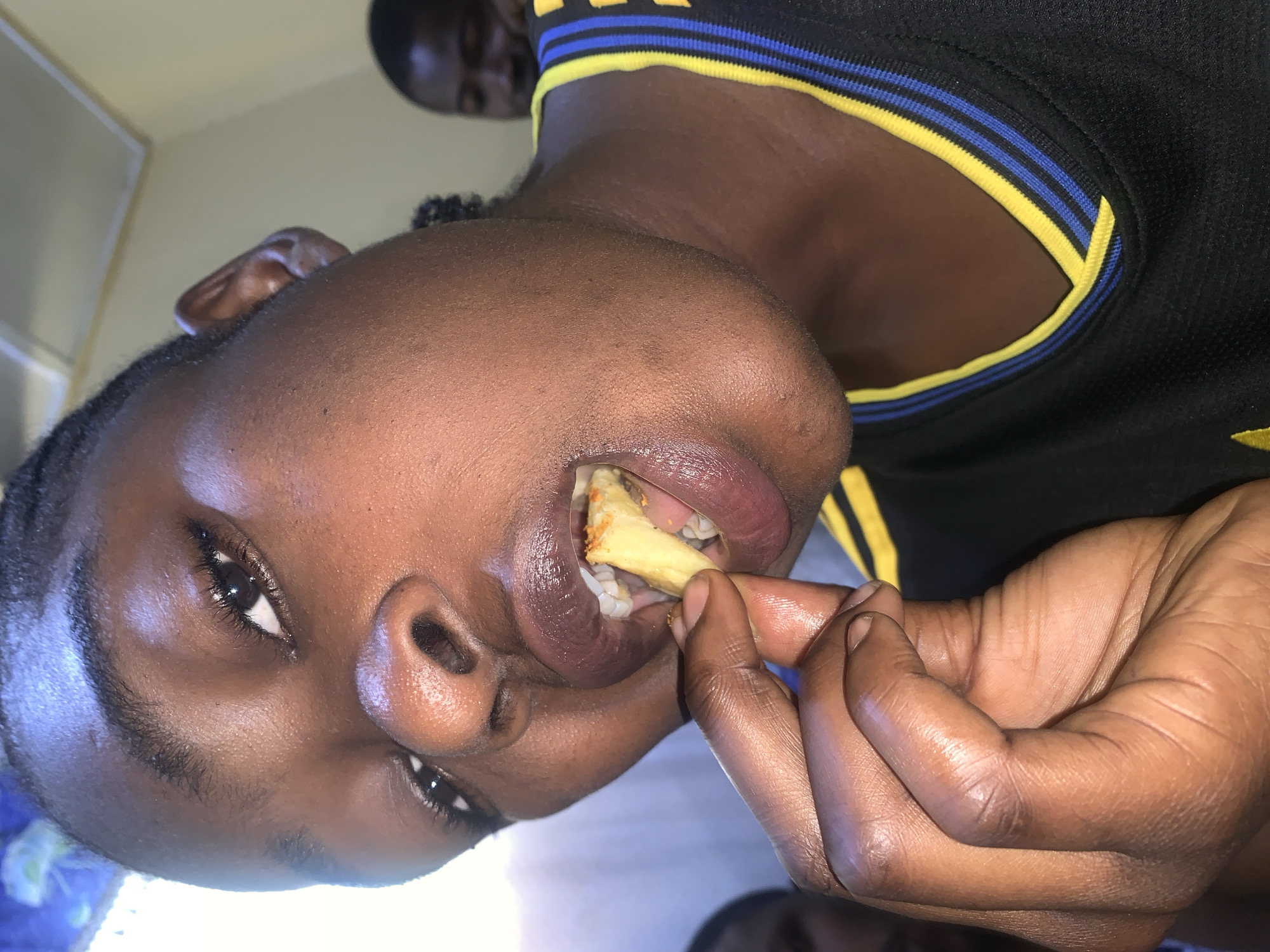 A close-up shot shows a person's face and hand as they eat a french fry. The person's mouth is open, and the french fry is partially inside their mouth. Their hand is holding the french fry up to their mouth. The person is wearing a black shirt with yellow and blue trim. In the background, another person is visible, out of focus. The scene appears to be indoors, with a light-colored wall and ceiling visible. The image was taken in Rafin Par, Nigeria.