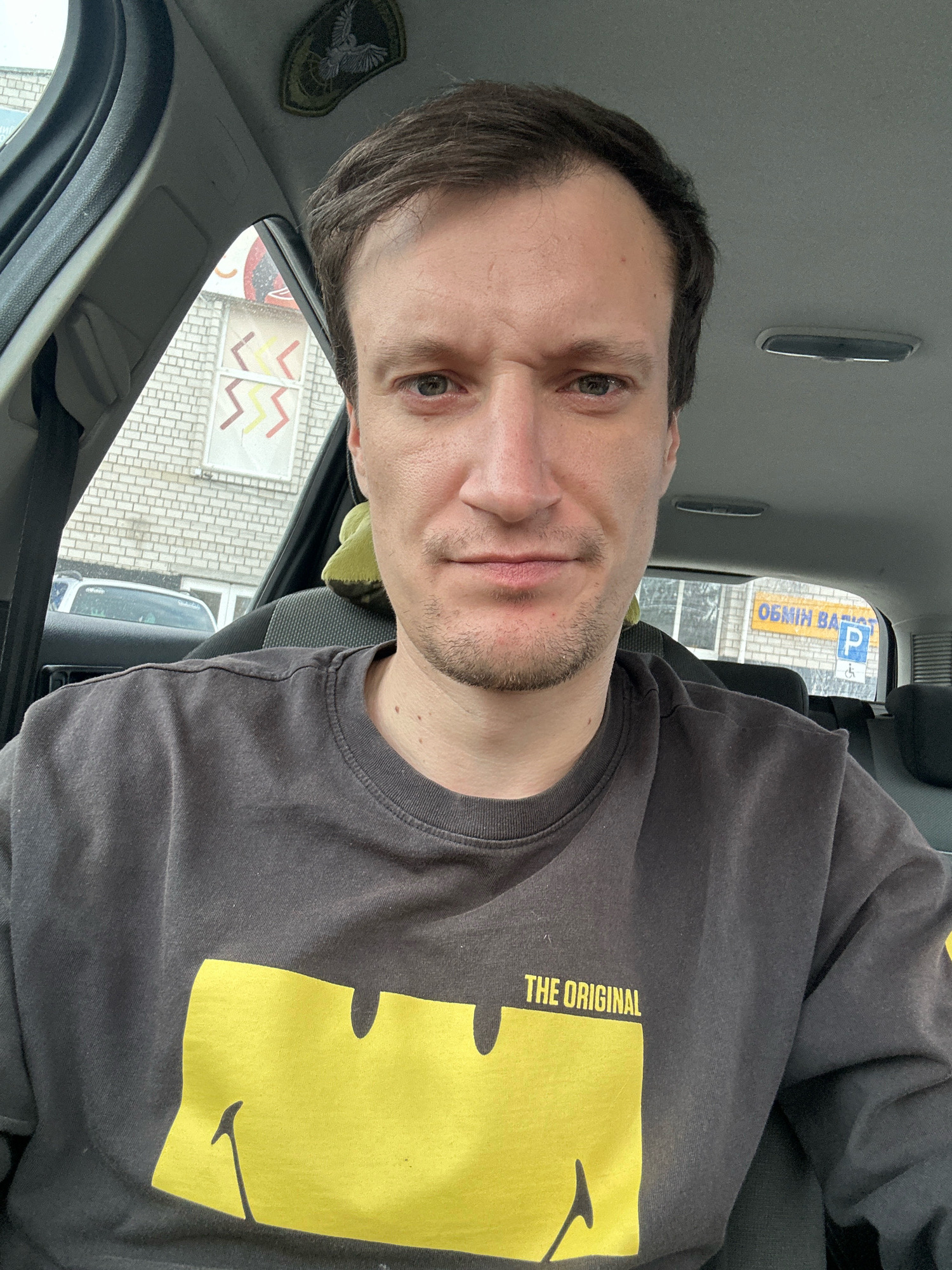 A man is photographed from the chest up, inside a vehicle. He is wearing a dark gray long-sleeved shirt with a large yellow graphic and the text "THE ORIGINAL" printed on it. The background includes part of the car's interior, a window revealing a brick building with some decorative elements, and a yellow sign with blue lettering and a blue parking symbol. The man has light skin, brown hair, and is looking directly at the camera with a neutral expression. The lighting appears to be natural daylight.