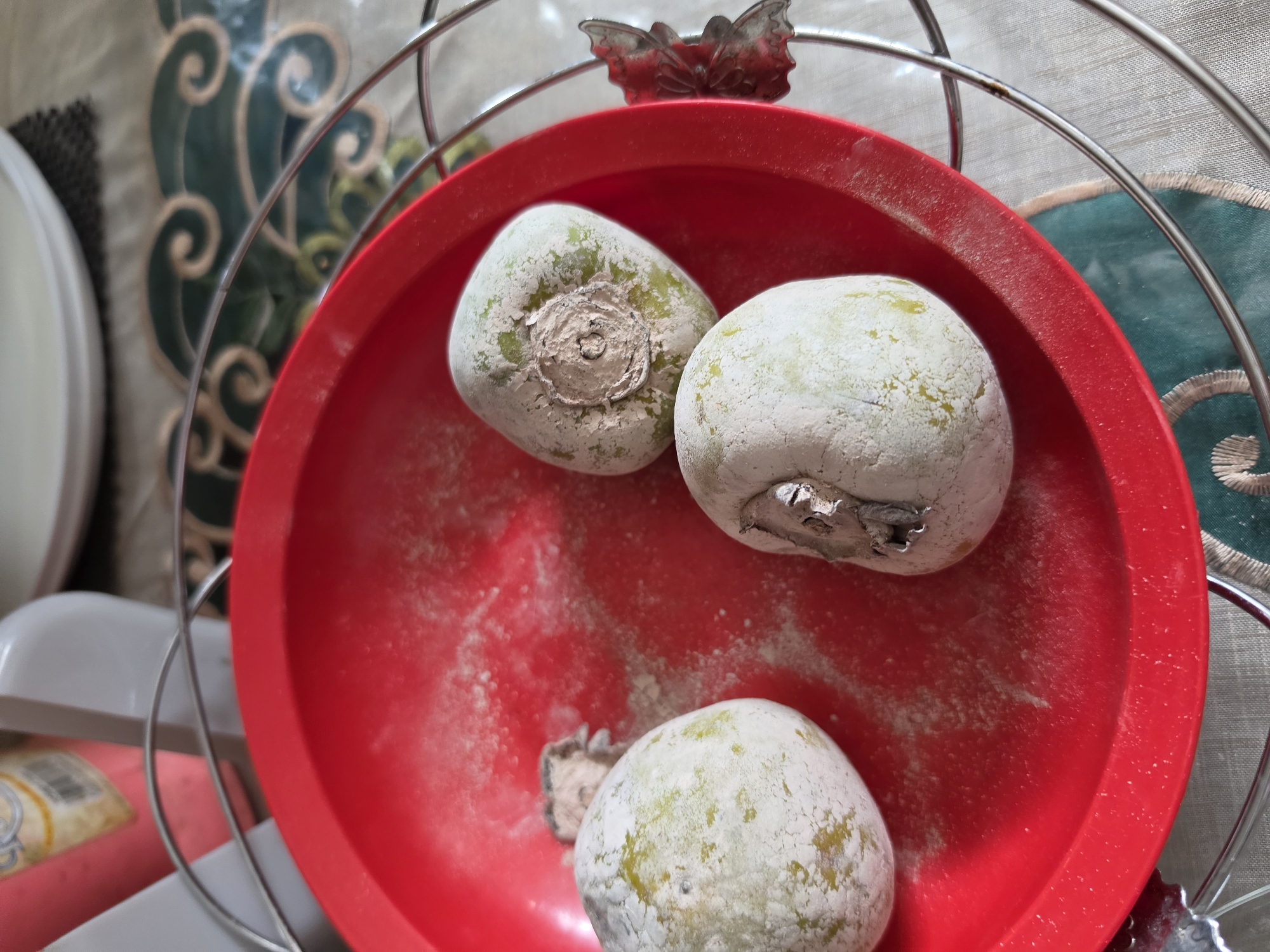A close-up shot shows three round, pale green fruits, possibly persimmons, in a bright red bowl. The fruits are covered in a powdery white residue and have visible stem scars. The bowl sits on a patterned tablecloth with green and white swirling designs. A silver wire rack surrounds the bowl, with a decorative metallic accent resembling a leaf or flower on the right side. In the background, out of focus, are bottles and containers, one pink with a yellow label. The lighting suggests an indoor setting. There is no indication of the specific location within the city of Medan, Indonesia.