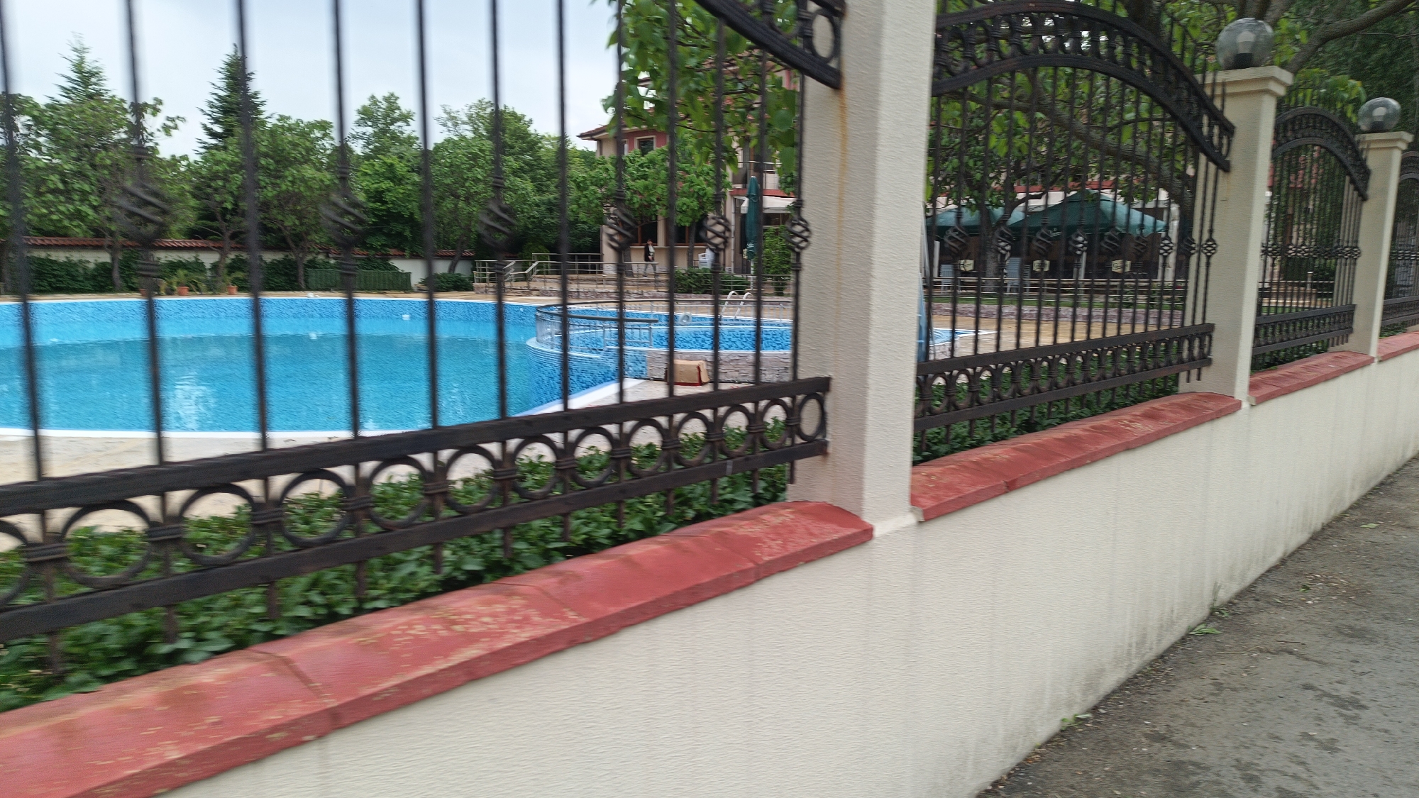The image shows an outdoor swimming pool partially visible through a decorative black metal fence with a repetitive circular pattern. The fence is mounted on a low white concrete wall topped with a red brick coping. Green shrubs are growing between the fence and the wall. The pool itself is lined with blue tiles and is surrounded by trees and buildings, partially obscured by the fence. The location is identified as Unknown, Unknown.